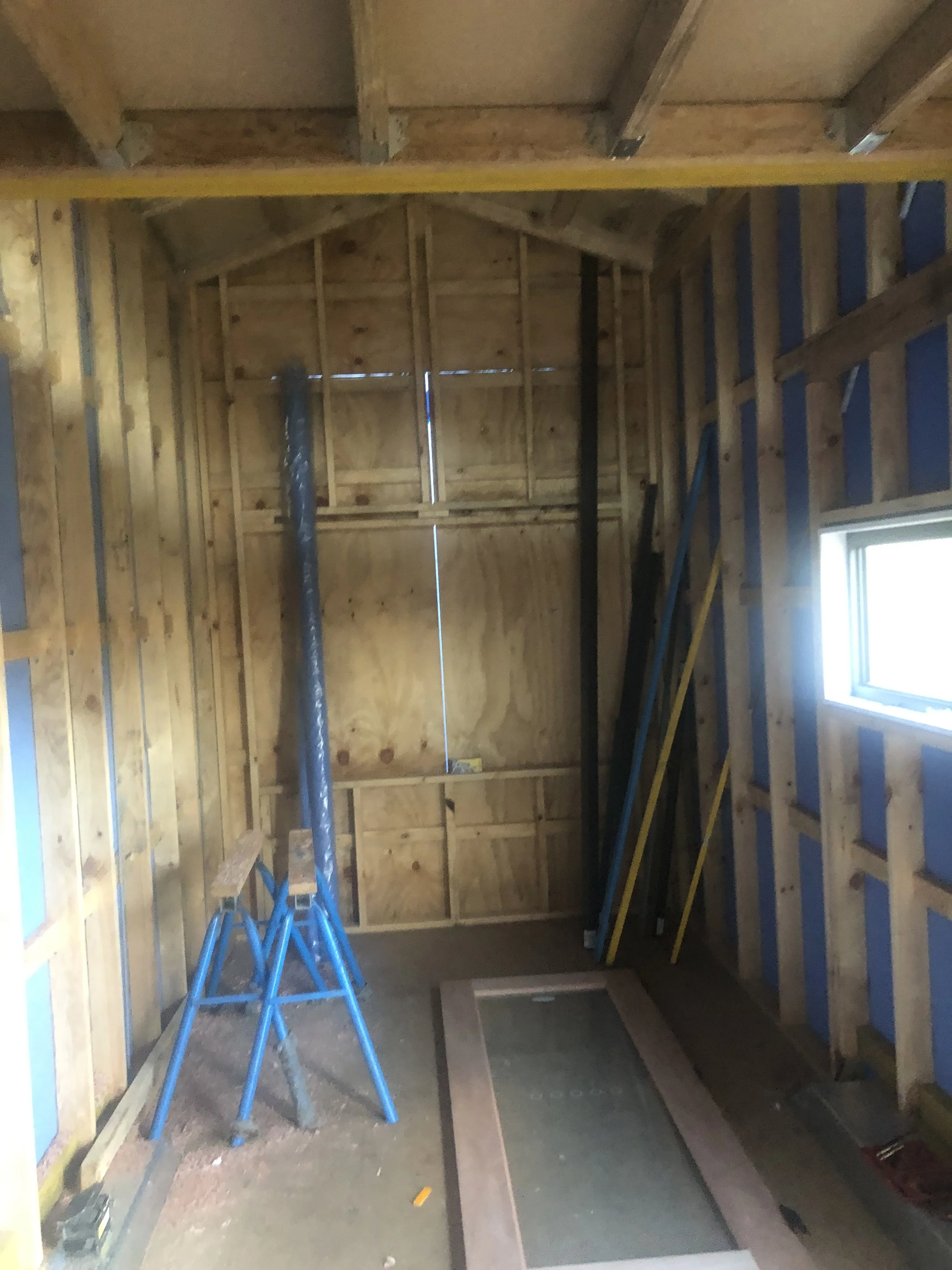 Interior of a building under construction with bare wooden framing, a small window on the right, and construction tools and materials, including a blue step ladder, a pole, and some framing boards.
