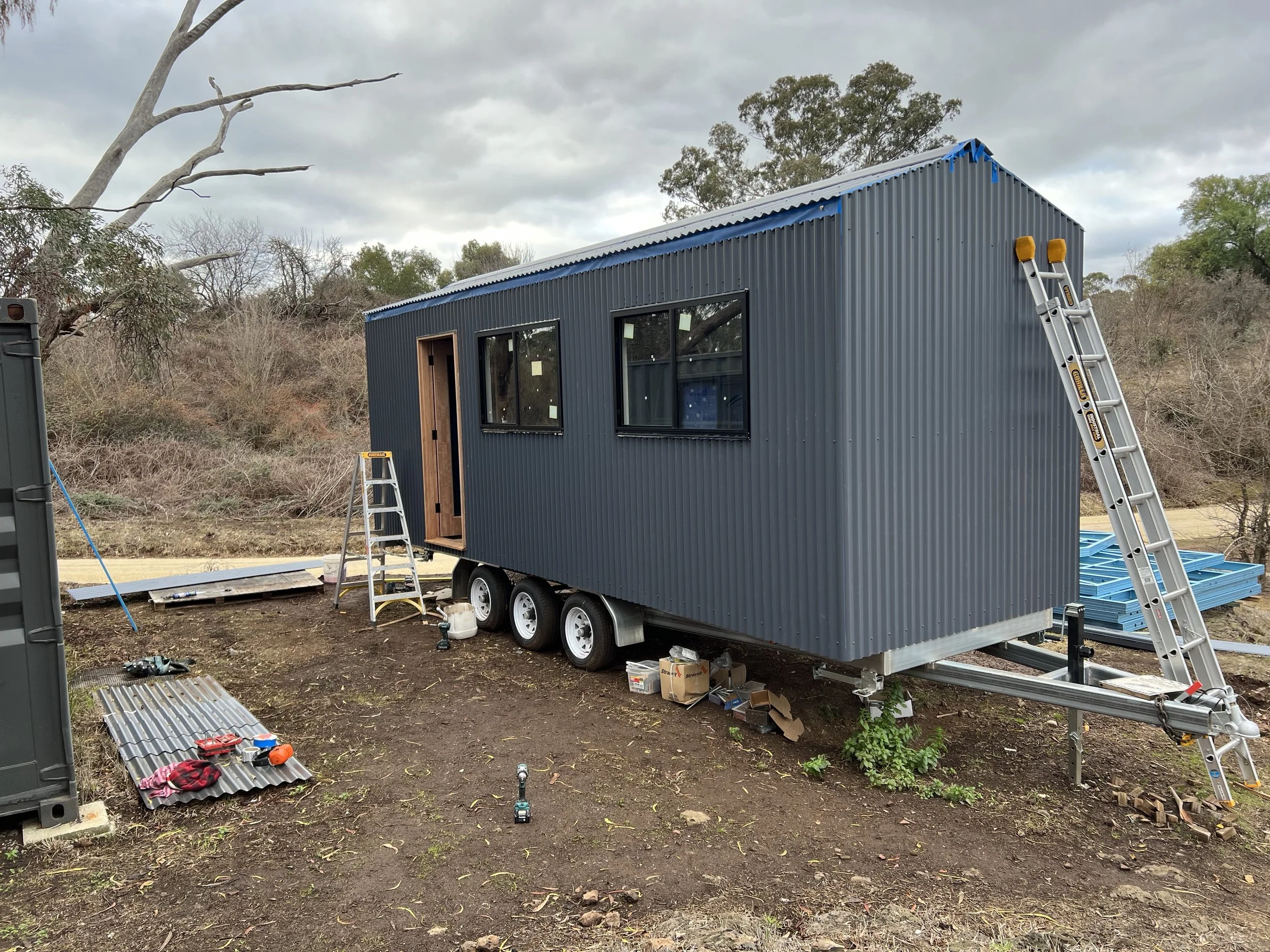 A tiny house under construction, mounted on a trailer with wheels, next to a ladder, tools, and building materials in an outdoor setting with trees and cloudy sky in the background.