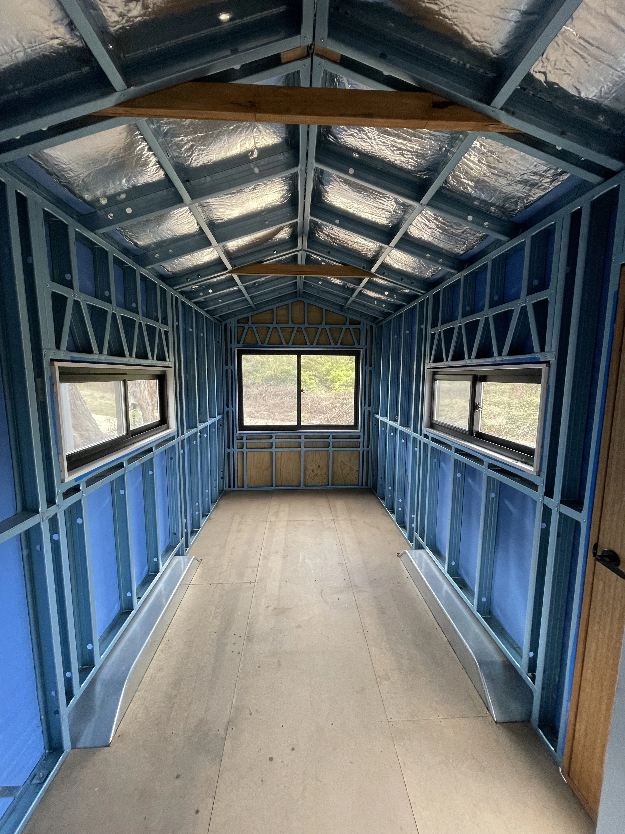 Interior view of a small room under construction with exposed blue metal framing, two horizontal sliding windows on each side, a large front window, silver metal window sills, wood ceiling beams, and a plywood floor.