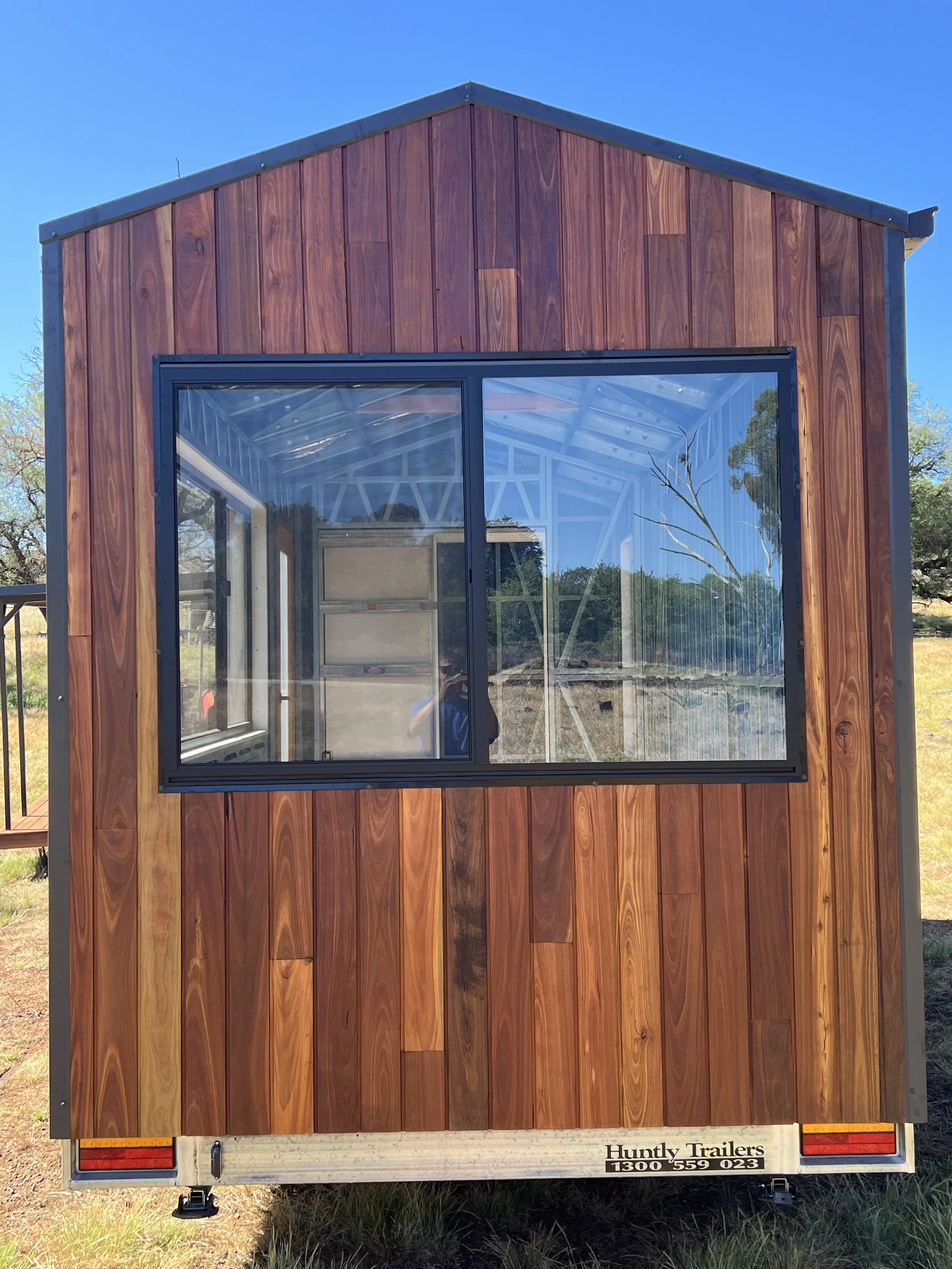 Rear view of a tiny house trailer with wooden siding and a large glass window, under a clear blue sky, with some trees in the background.