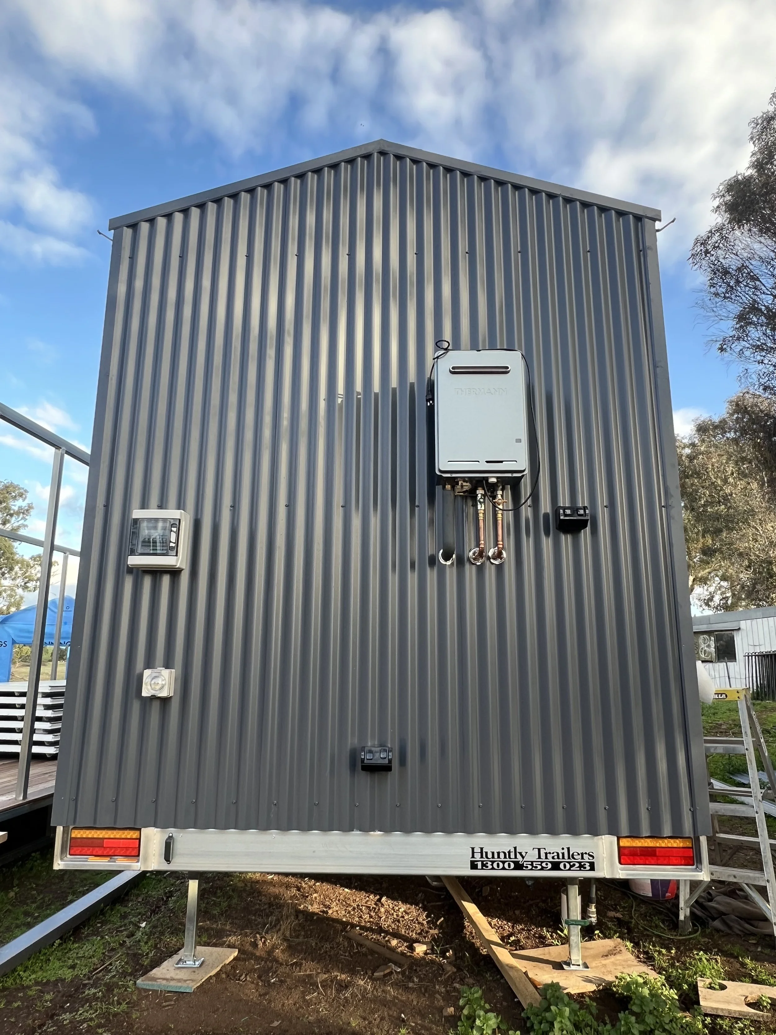 Gray metal trailer with electrical and mechanical equipment mounted on the back, labeled 'Huntly Trailers,' parked on a grassy area under a partly cloudy sky.