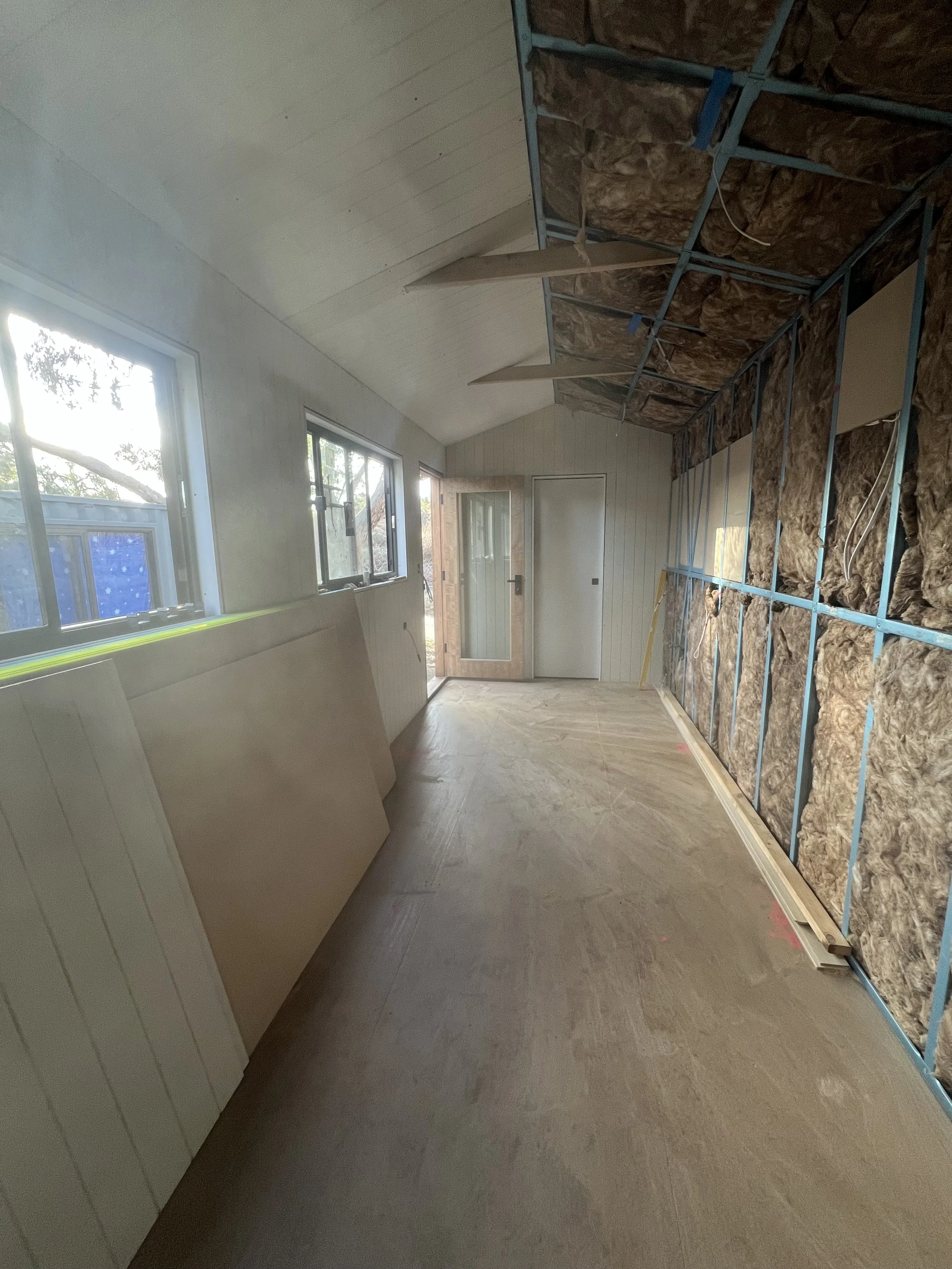 Interior of a room under construction with exposed wall insulation, partially installed ceiling panels, windows, and a door at the back.