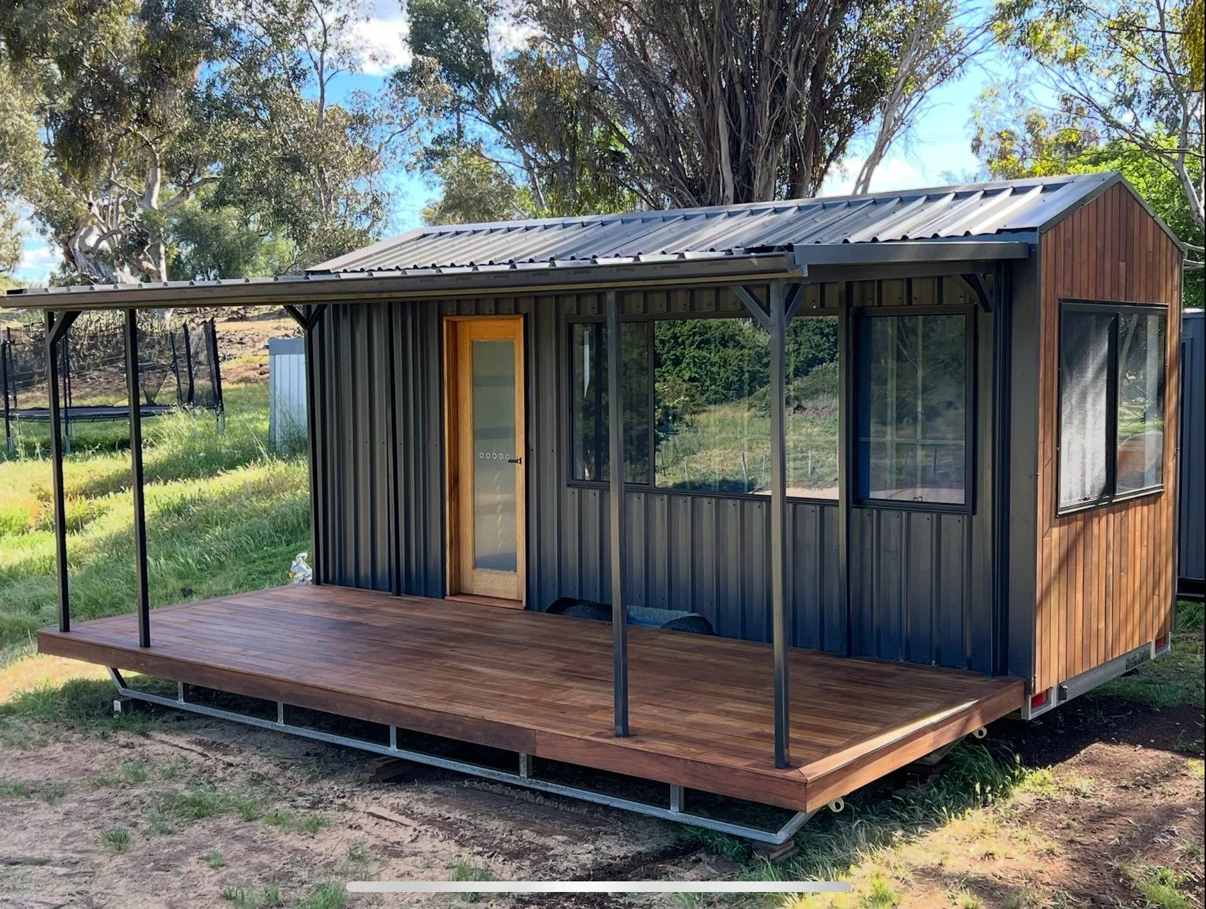 A small, portable house with a combination of metal and wood exterior, a metal roof, a wooden porch, and large windows, situated on a grassy area with trees in the background.