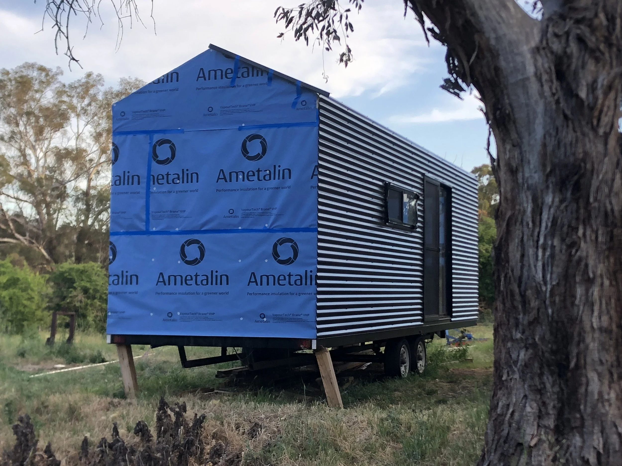 A small building under construction with blue insulation wrap labeled 'Ametalin' attached to the exterior, situated in a grassy, wooded area, with a tree trunk partially visible in the foreground.