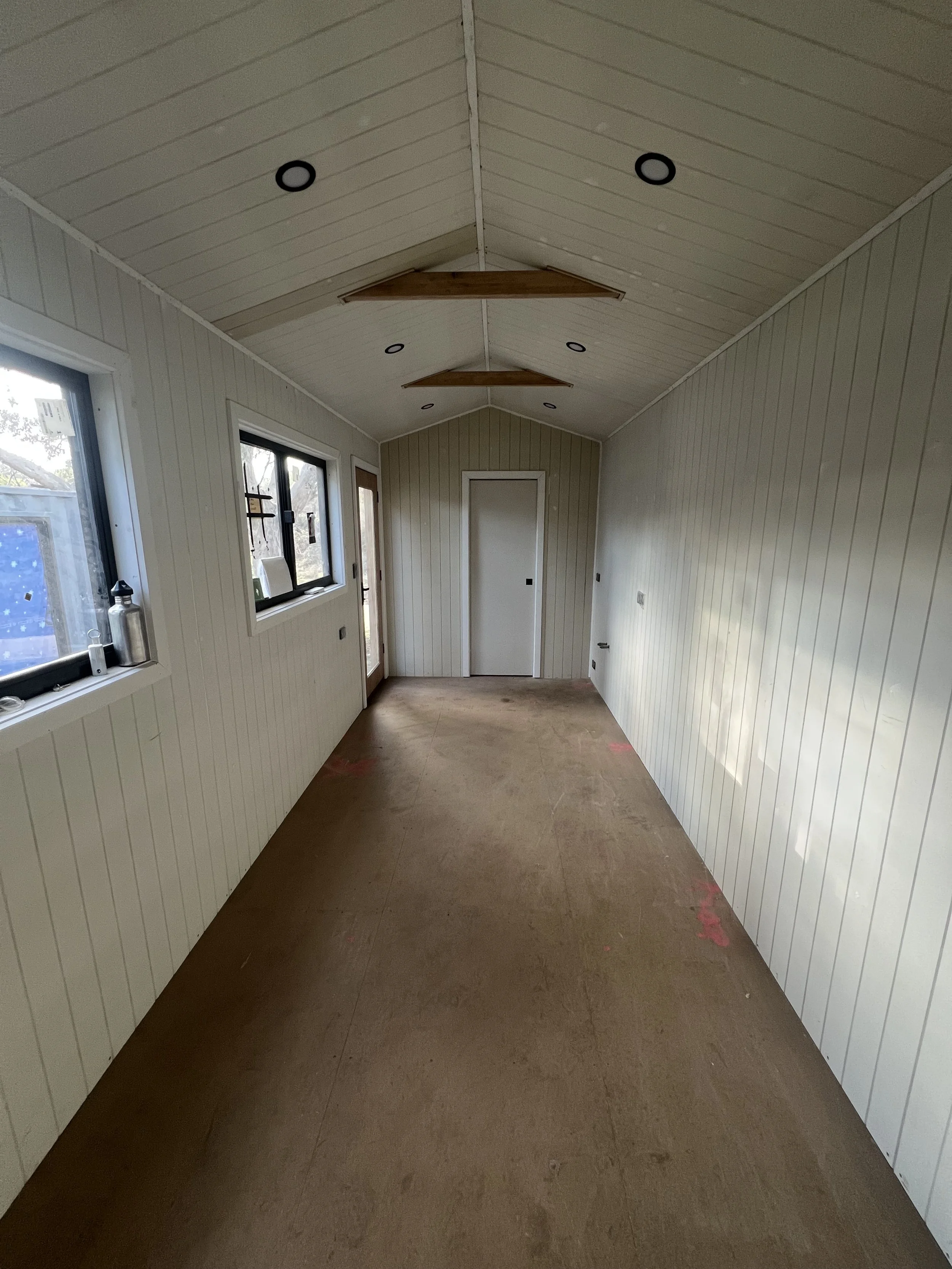 Empty room with white paneled walls and ceiling, two windows on the left, a door at the end, and a ceiling fan with wooden blades, under construction or renovation.