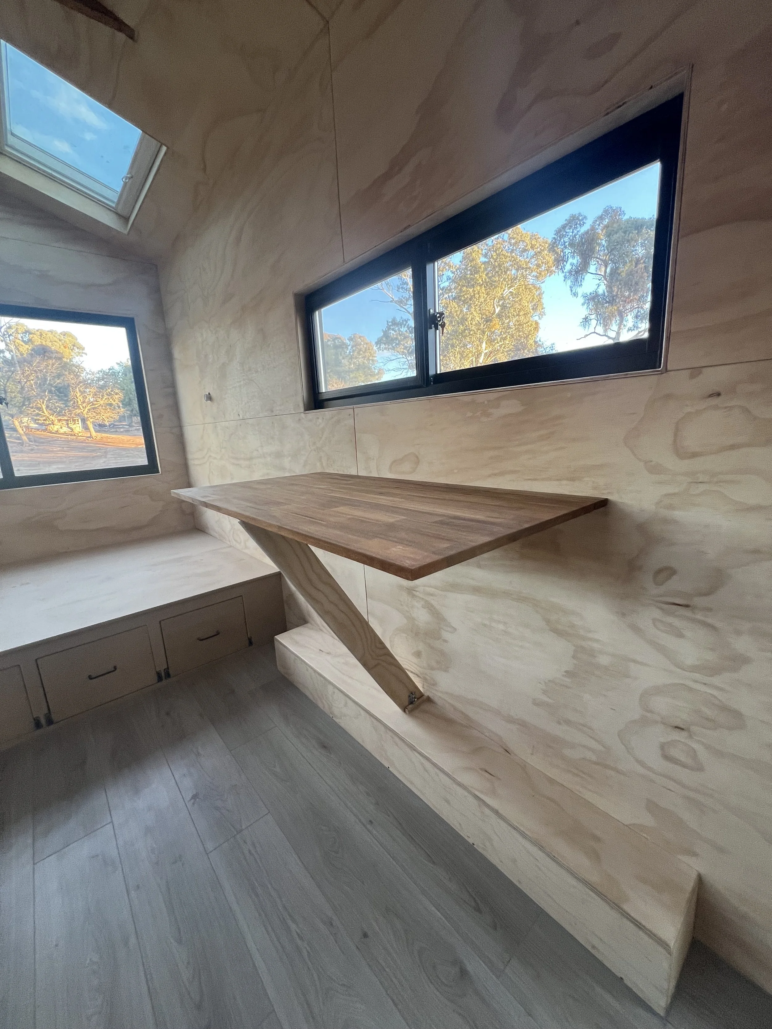 A small room with wooden walls and a wooden floor, featuring a rectangular window and a tilted wooden shelf attached to the wall, with a cubby storage area beneath the window.