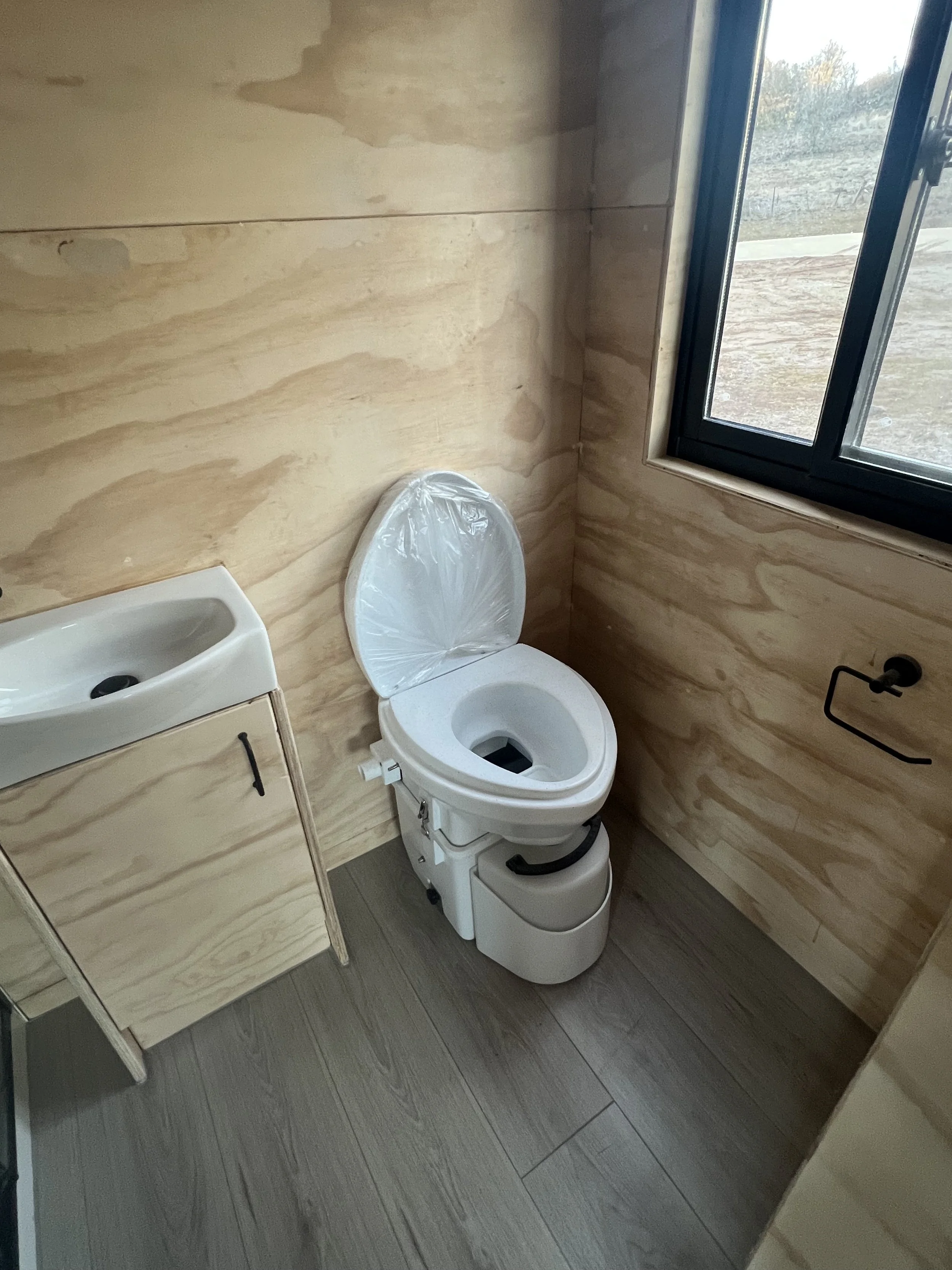 A small bathroom with a wooden wall, a window, a toilet with a plastic cover on the seat, a small sink with a cabinet, and a toilet paper holder.