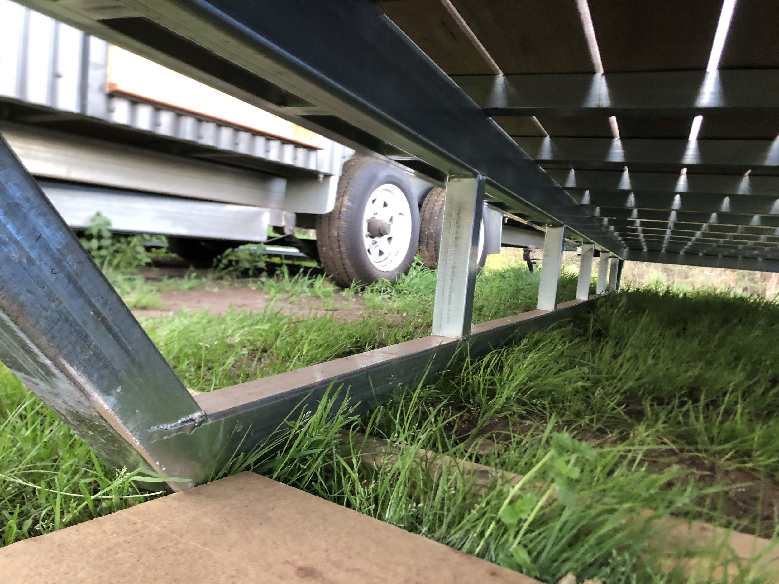 The underside of steel skid frame for deck supported by metal legs, with grass and a part of a vehicle with a tire visible in the background.