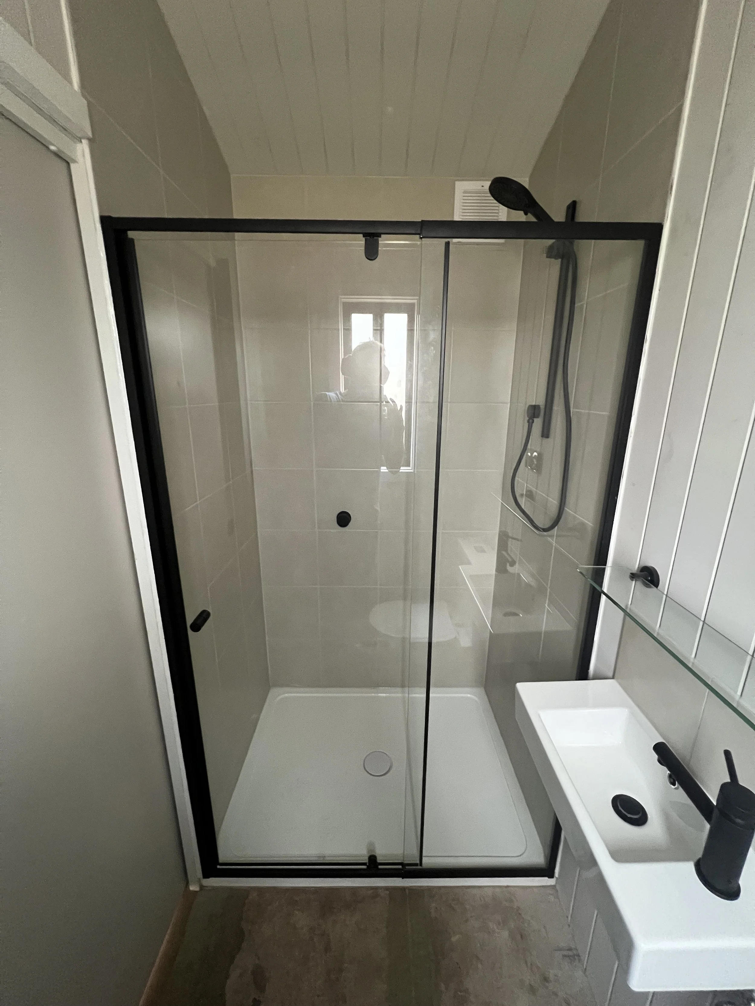 A modern bathroom featuring a walk-in shower with black framing and clear glass doors, a small window on the back wall, a handheld showerhead, a compact white sink with a black faucet, and neutral-colored wall tiles.