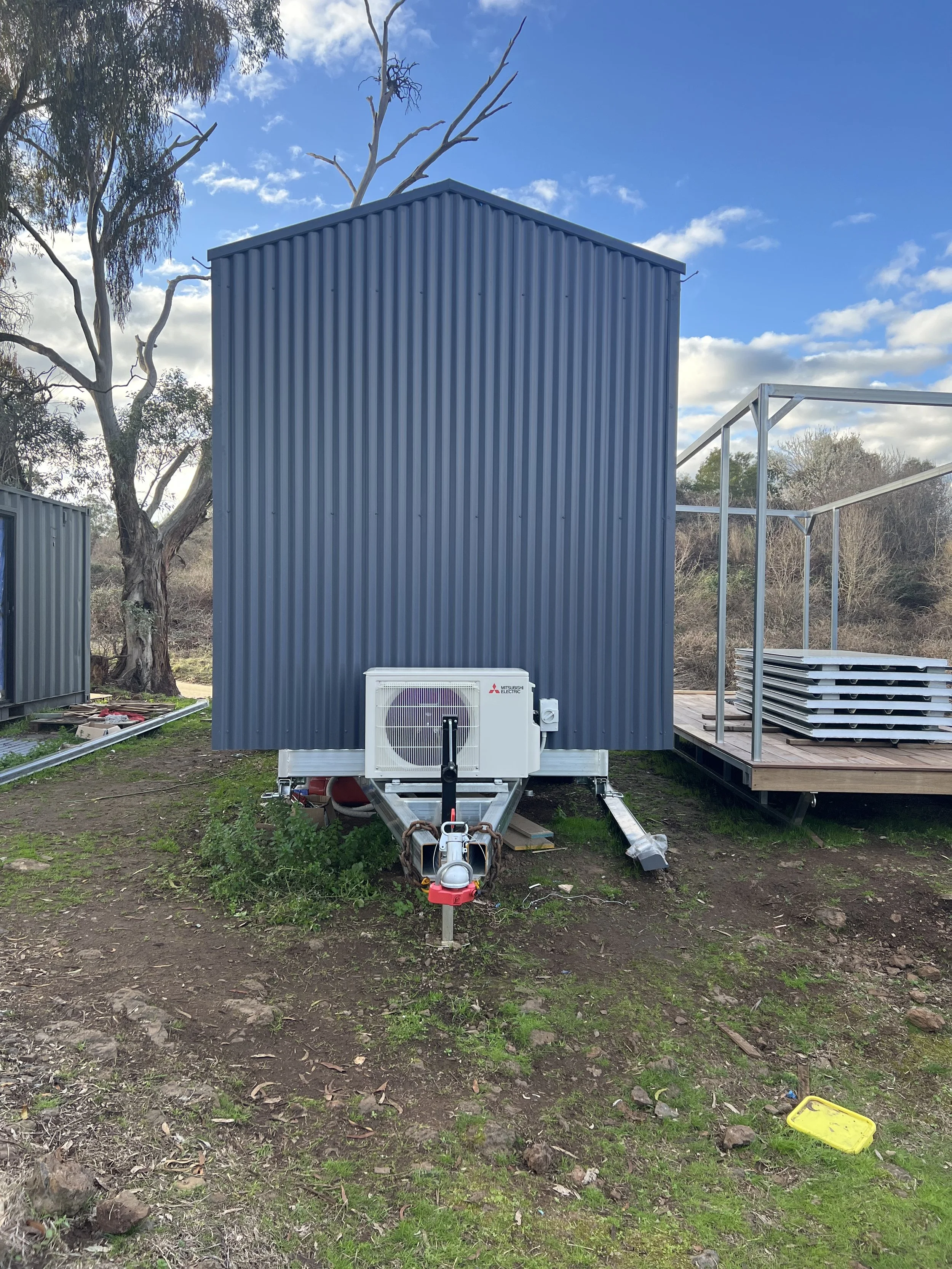 Blue metal tiny house with a Mitsubishi mini split air conditioning unit mounted on the front, situated on a grassy dirt patch with a tree and construction materials nearby.
