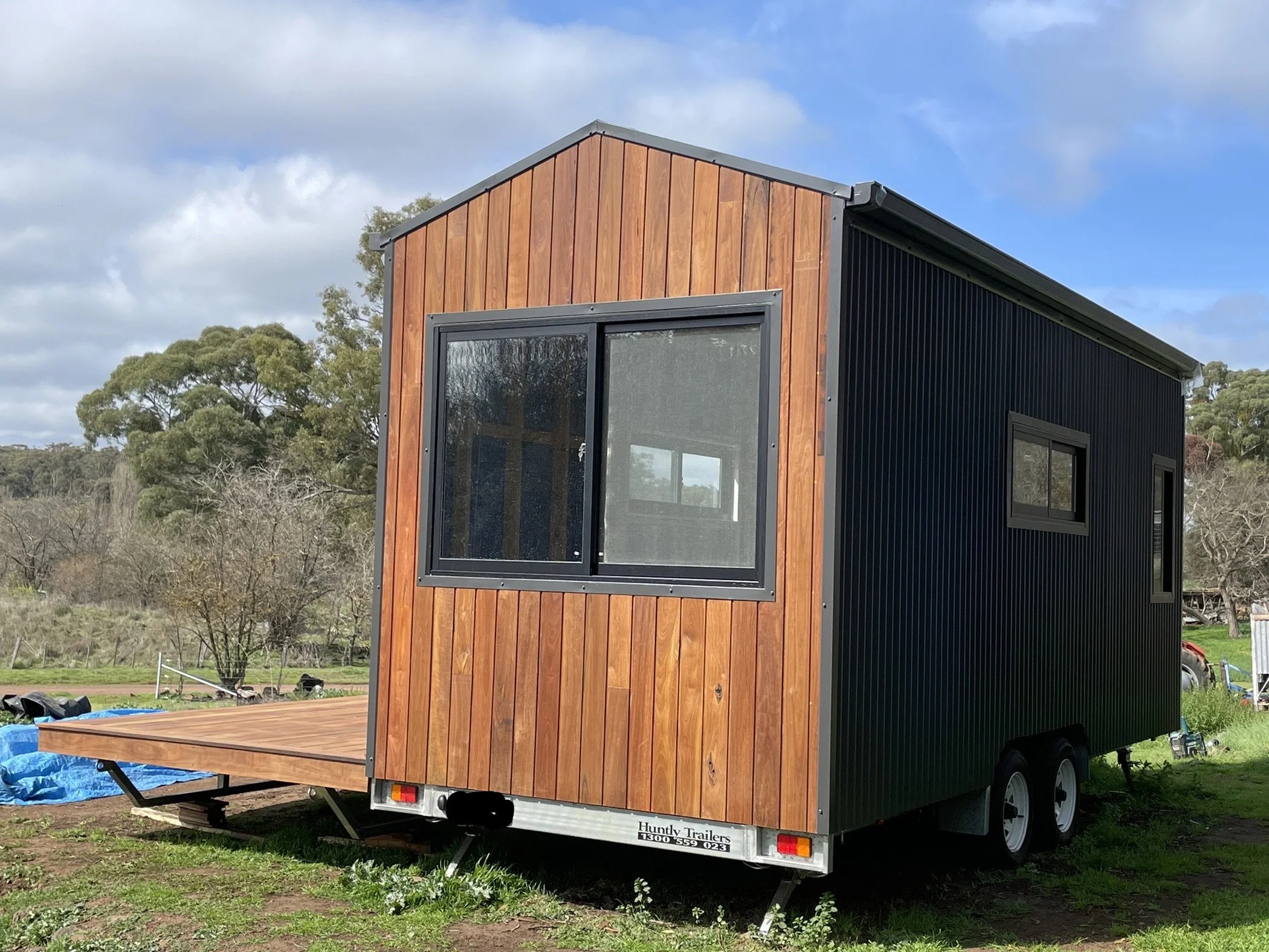 A modern tiny house trailer with a wooden exterior on one side and black corrugated metal on the other, parked on grass in a rural setting with trees and cloudy sky in the background.