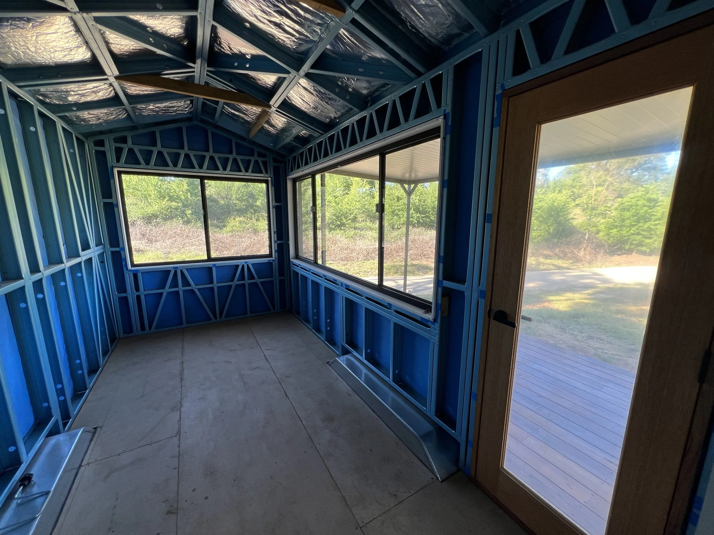Interior of a small building under construction with blue metal framing, two large windows, and a door with a large glass panel leading outside to a deck.