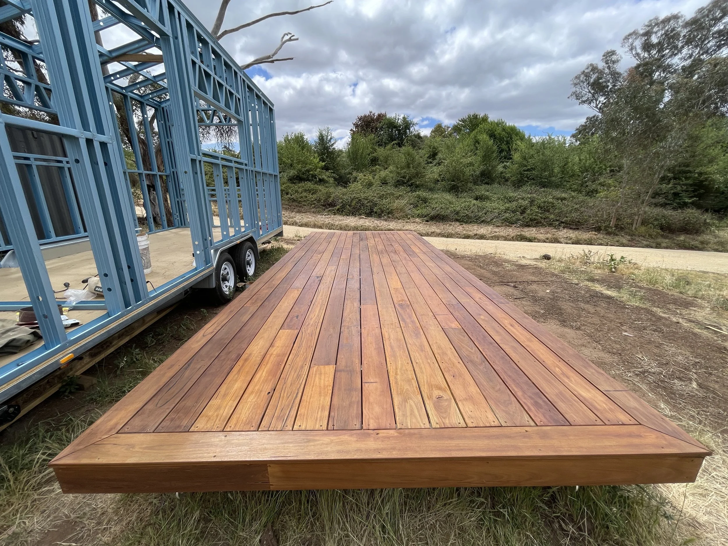 A freshly constructed wooden deck outdoors with a blue trailer nearby under a cloudy sky, surrounded by greenery and trees.