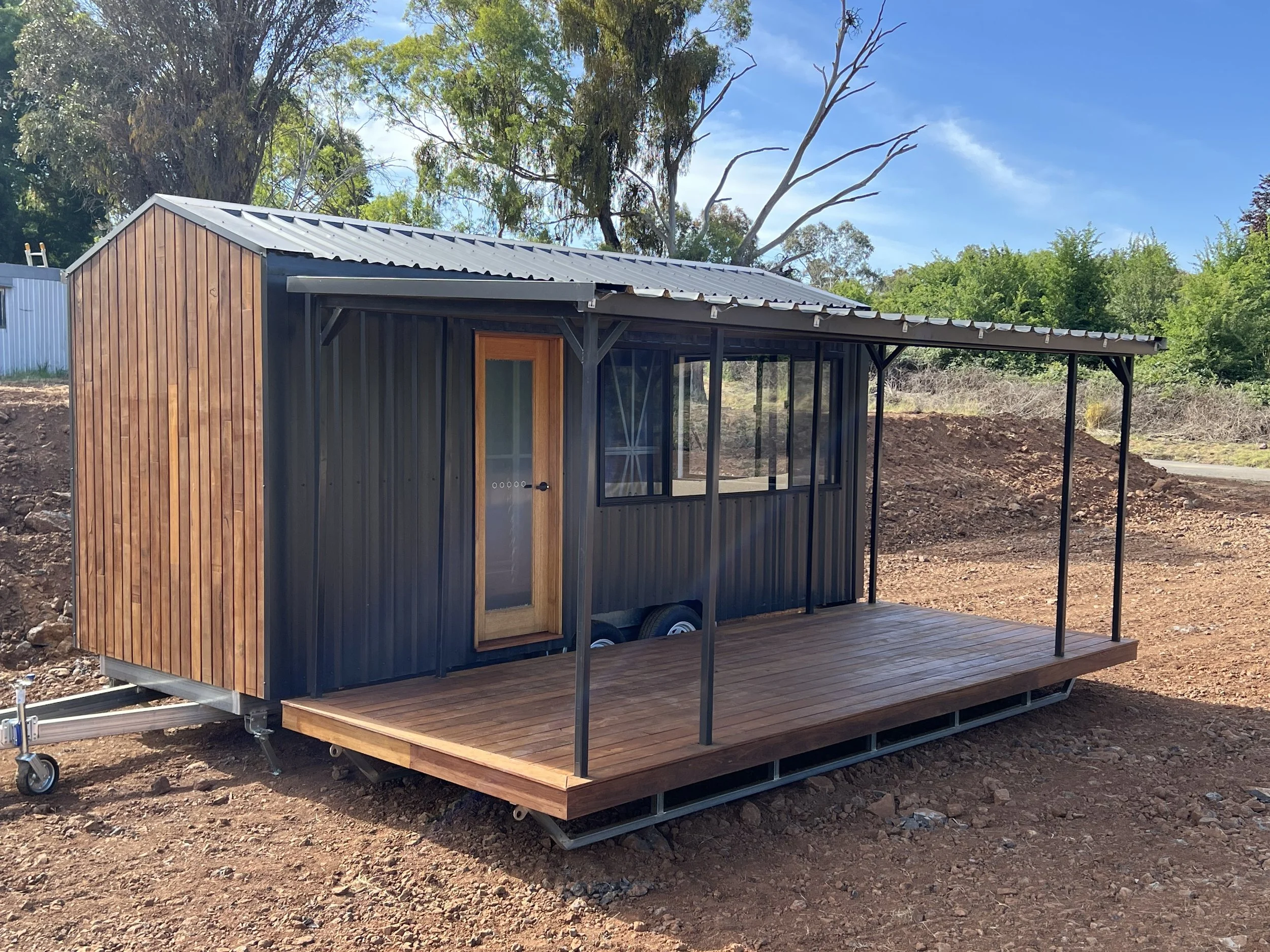 Small modern tiny house with monument trimdek cladding, a wooden door, and a small wooden porch, set on a cleared outdoor area with trees in the background.