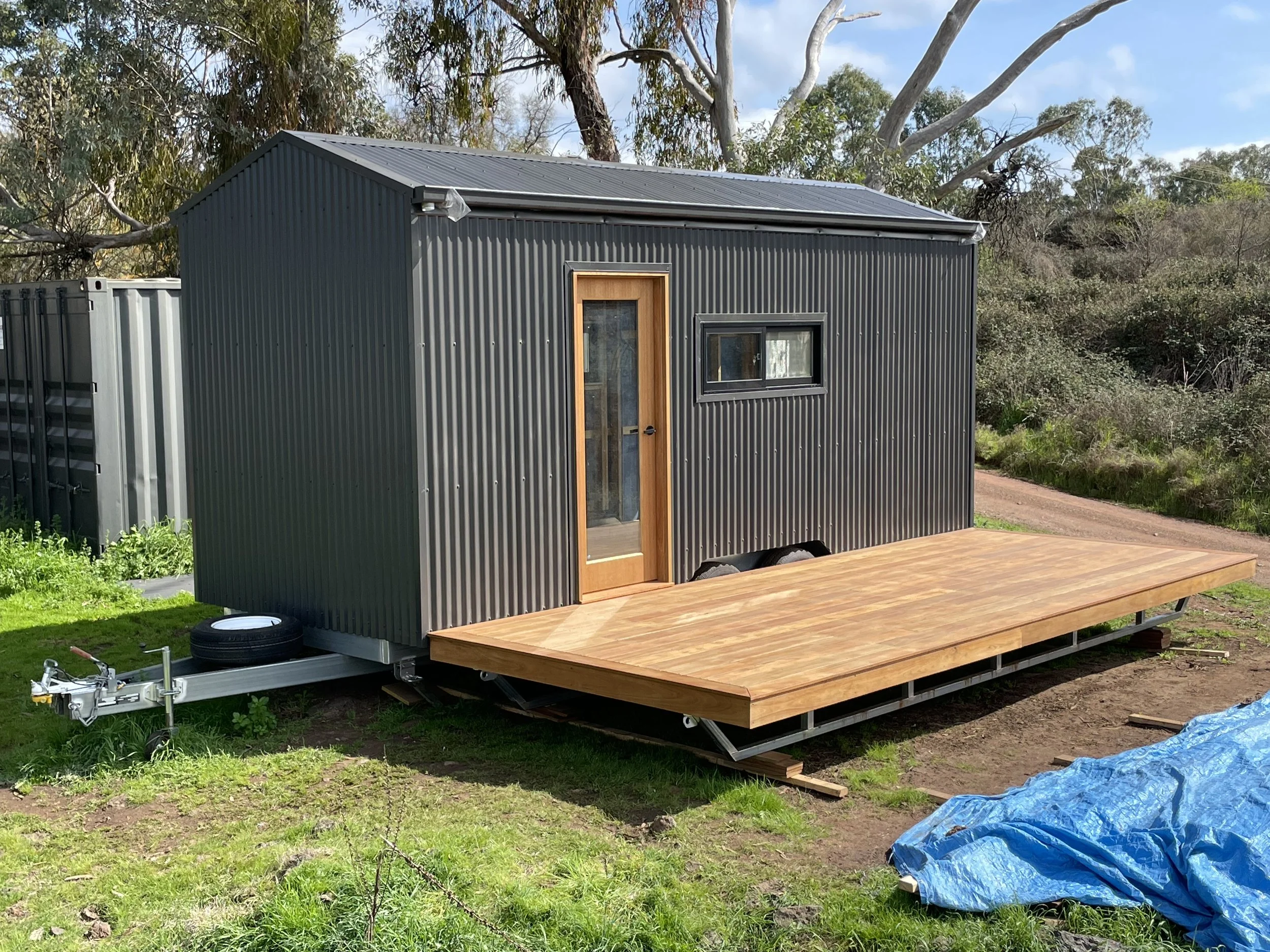 A newly constructed tiny house with dark metal siding and a wooden front door, built on a trailer with a wooden deck extension. The house is situated outdoors on grassy terrain with trees and a dirt path in the background.