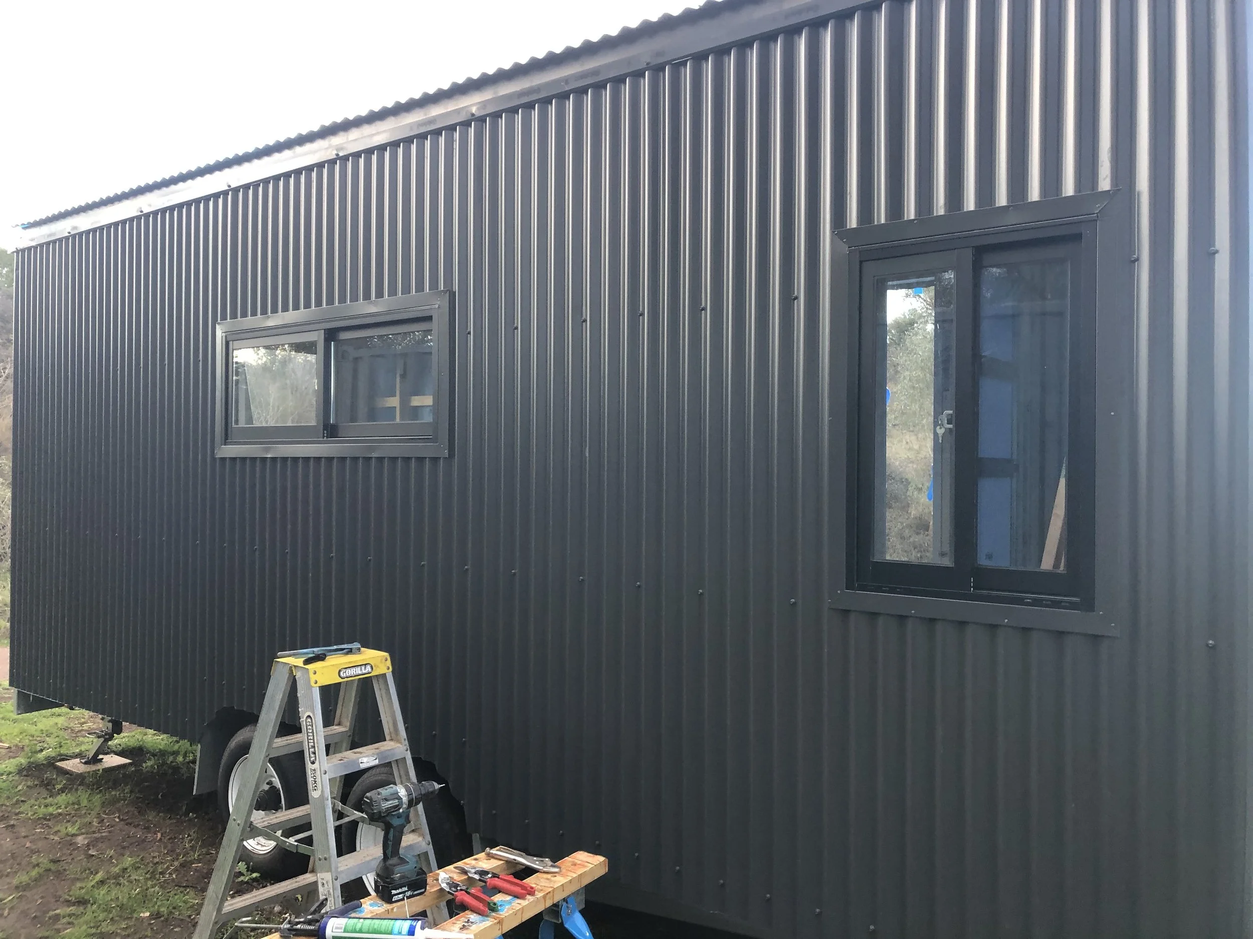 A tiny house with monument custom orb cladding under construction with two windows, a ladder, and construction tools outside on the ground.