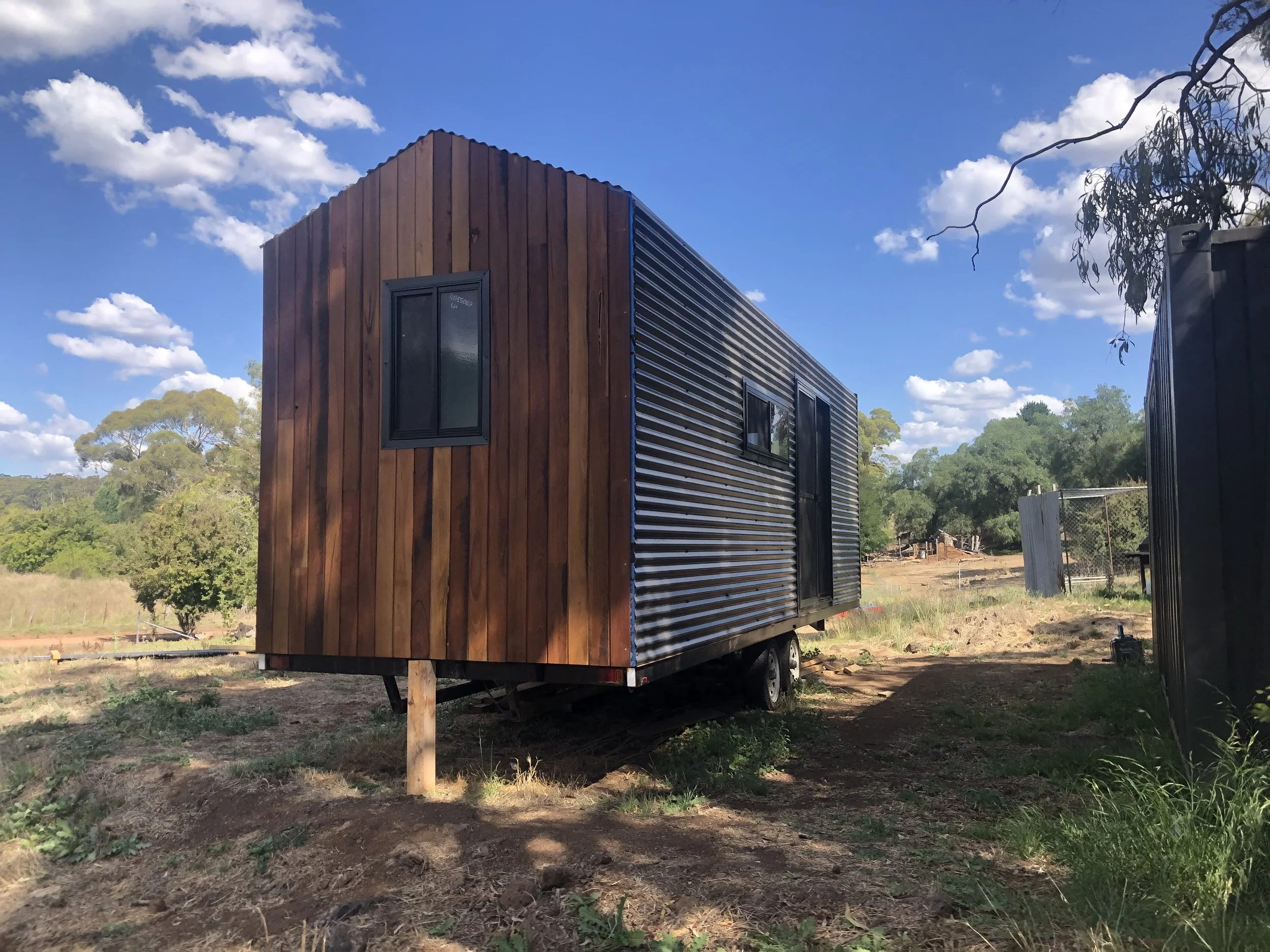 A tiny house on wheels, with a wooden exterior on the front and corrugated metal siding, on a dirt lot under a partly cloudy sky.
