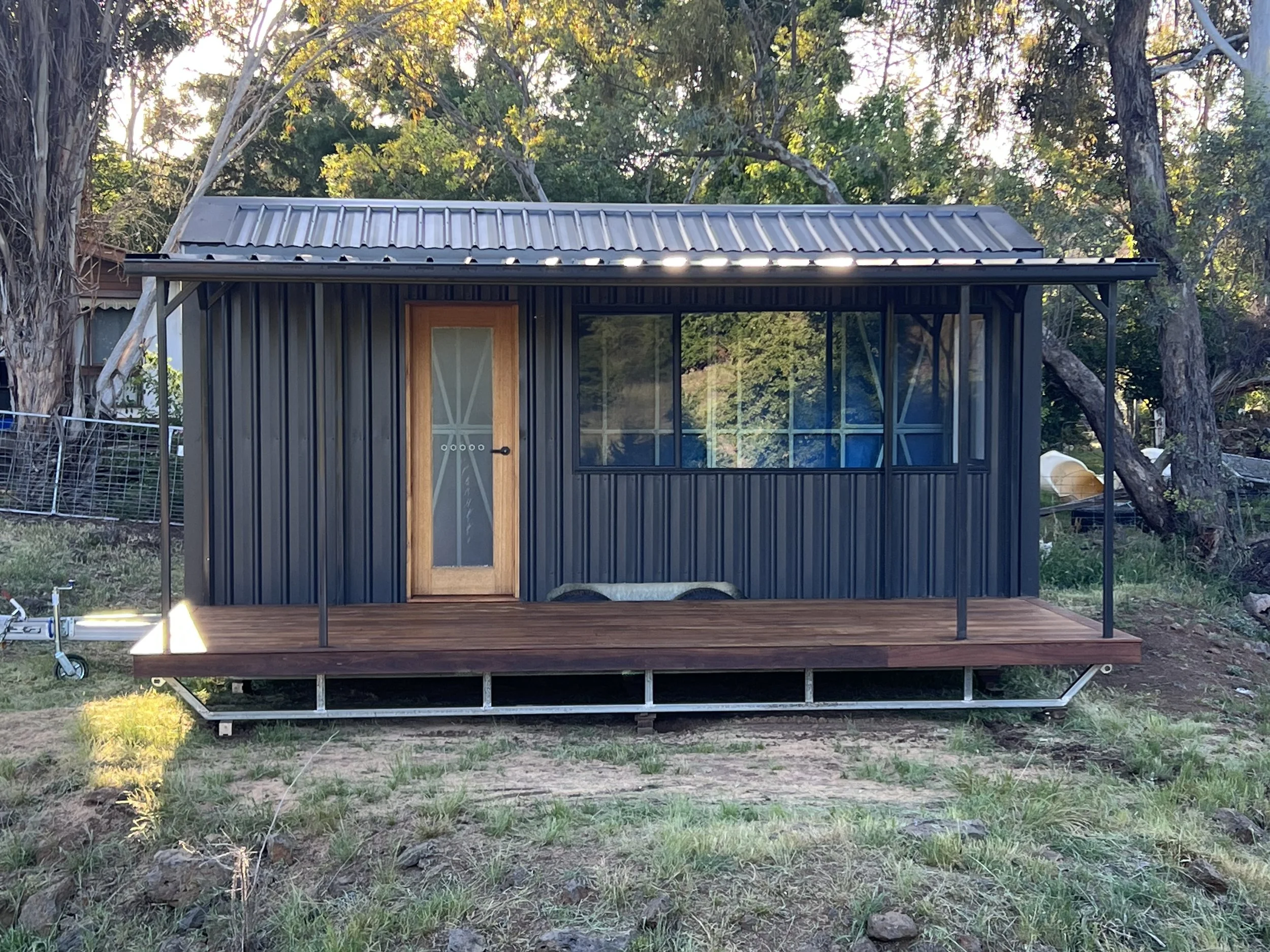 A small modern house with black metal siding, a wooden door with glass panels, and a screened front porch with a wooden deck, situated outdoors among trees and grass.