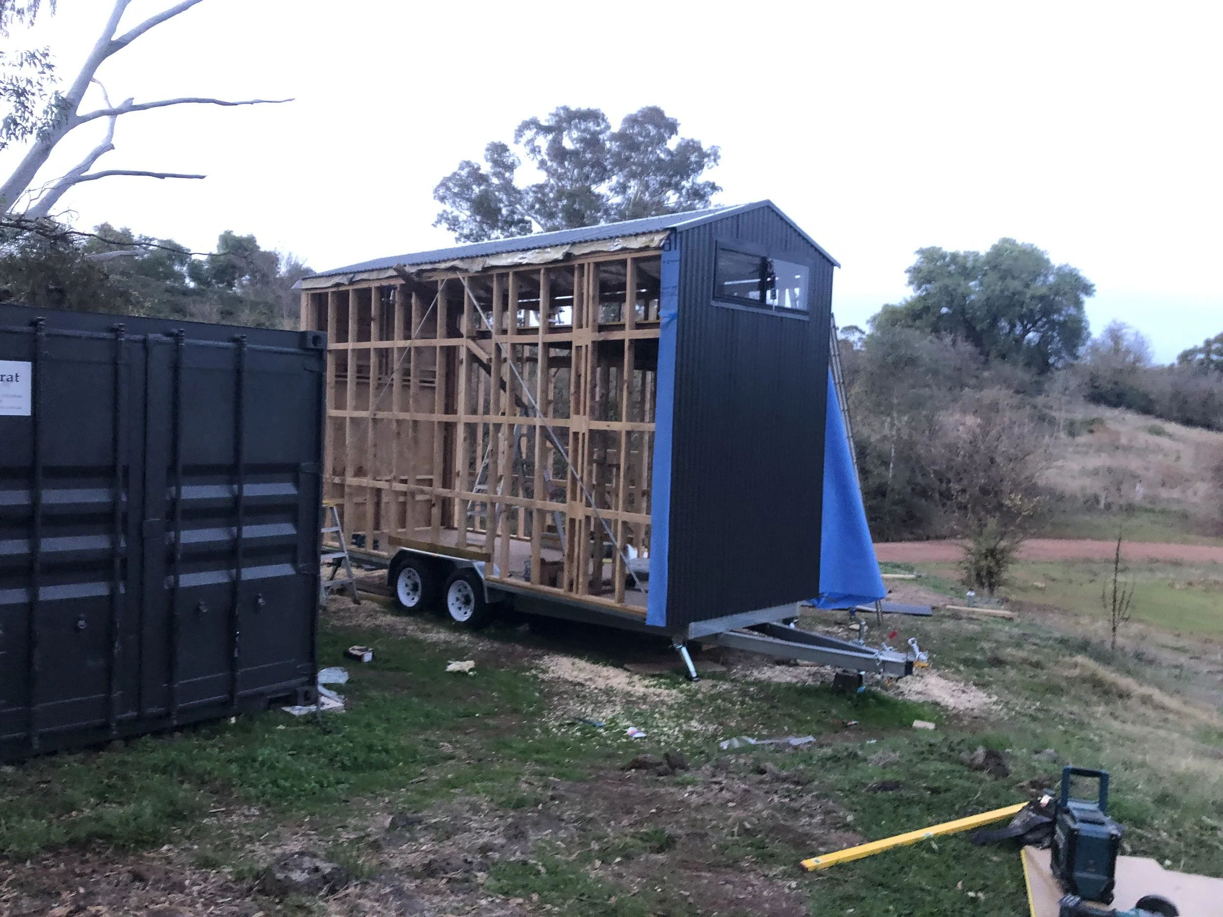 A small mobile home under construction on a trailer, with a partially completed wooden frame and a dark blue exterior panel, set in a rural outdoor area with trees and grassy hills in the background.