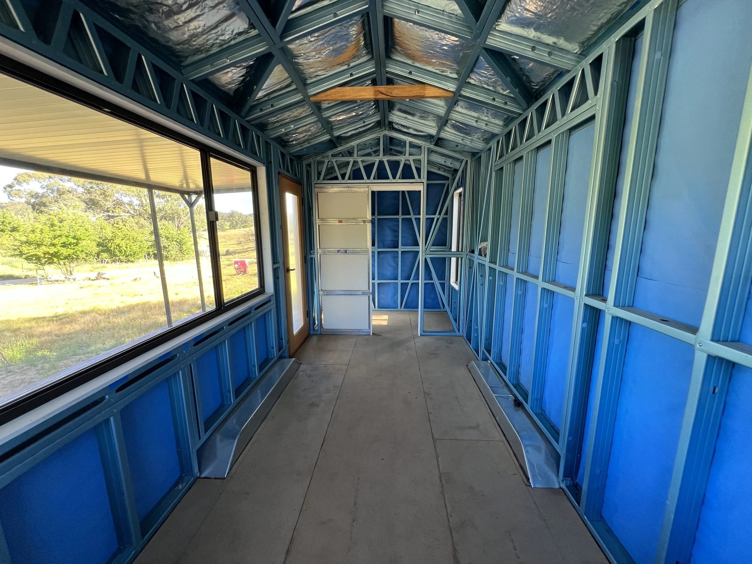 Interior of a partially finished building with blue metal framing, large windows on one side, and a concrete floor. Outside, trees and grass are visible.