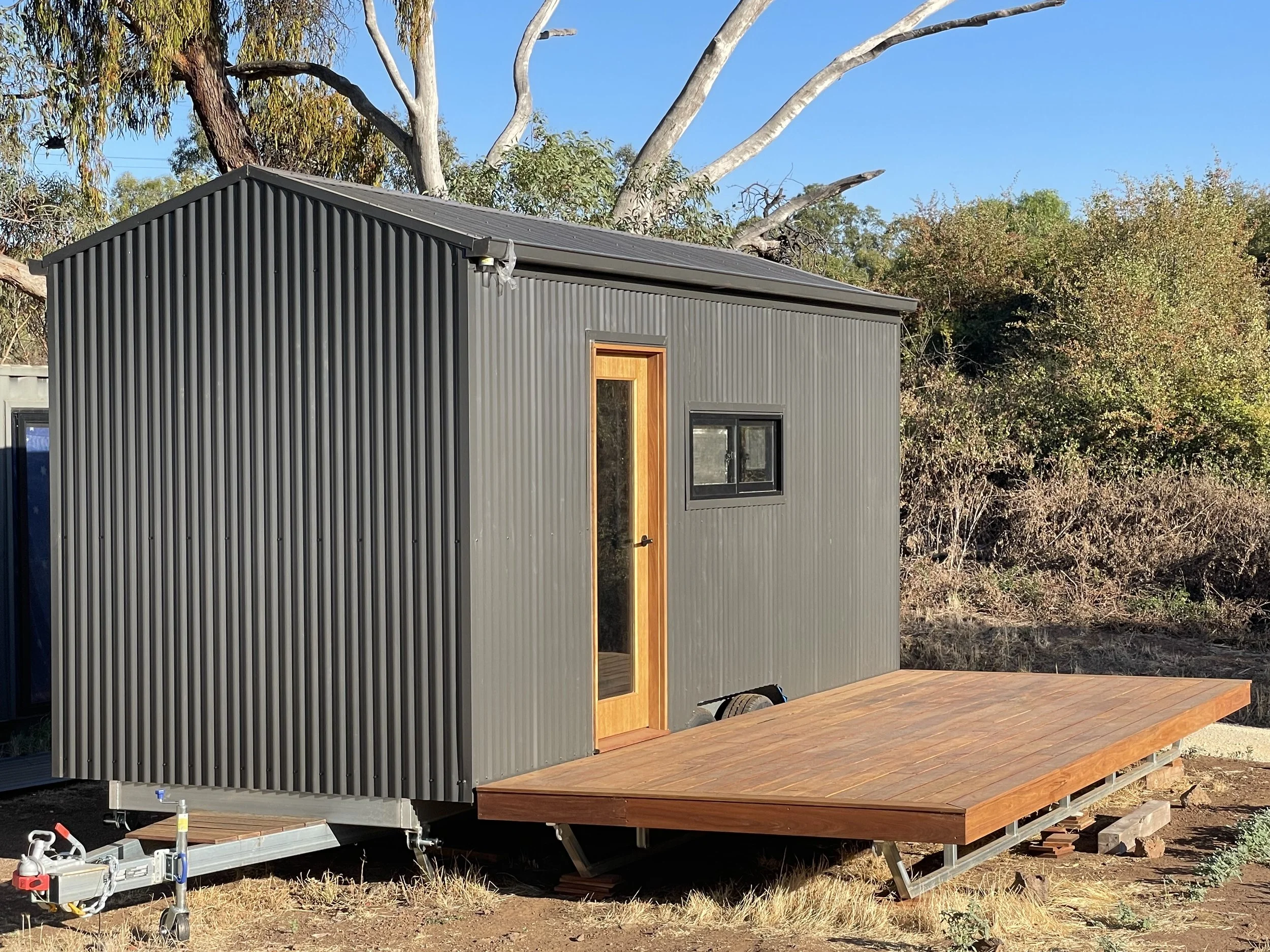 Small, modern tiny house with black corrugated metal exterior, a wooden door, and a small window, situated on a portable trailer with a wooden deck outside, in a natural outdoor setting with trees and clear blue sky.