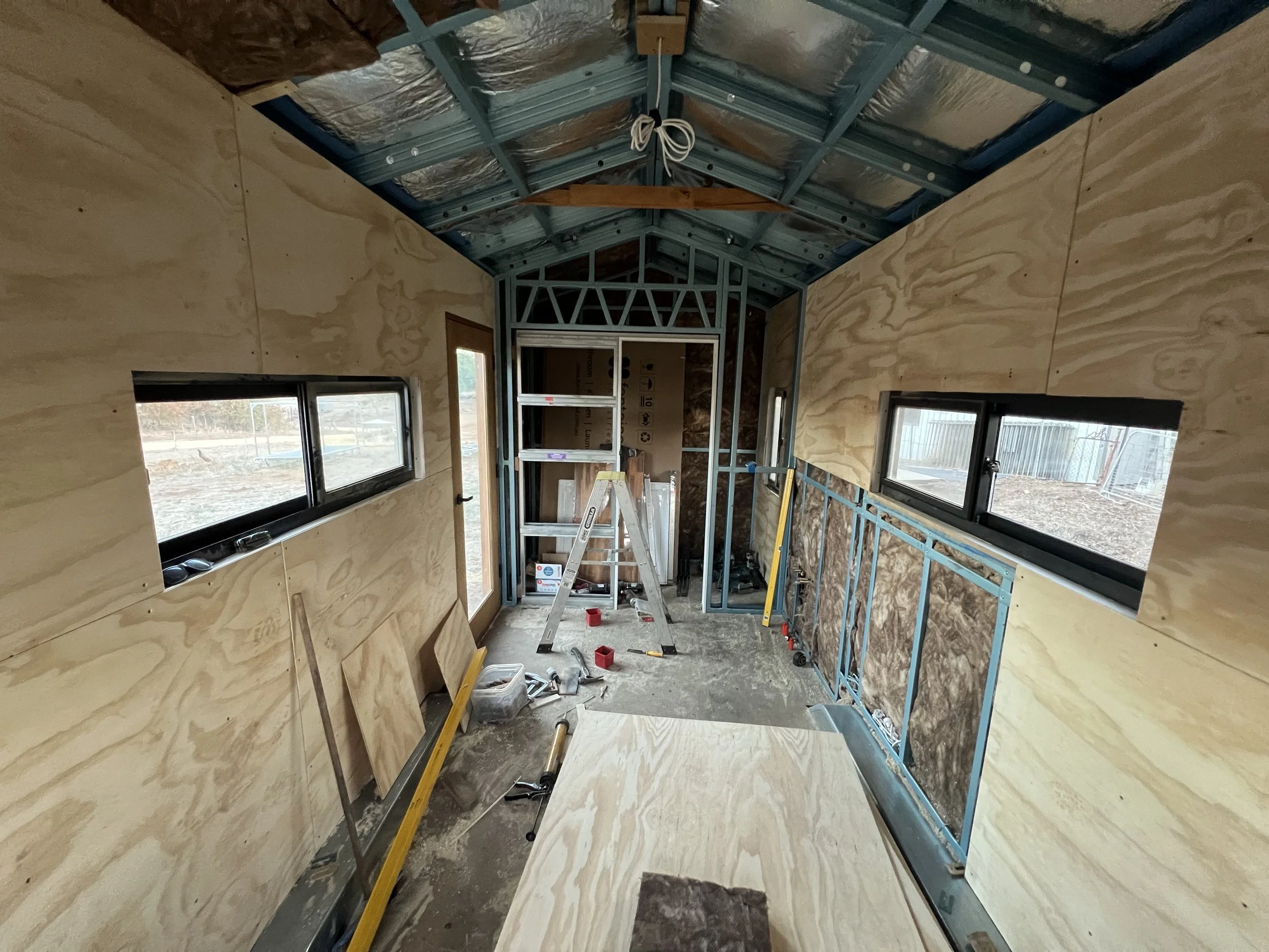 Interior of a building under construction with unfinished plywood walls, metal framing, and windows installed. Construction tools and materials are scattered on the floor.