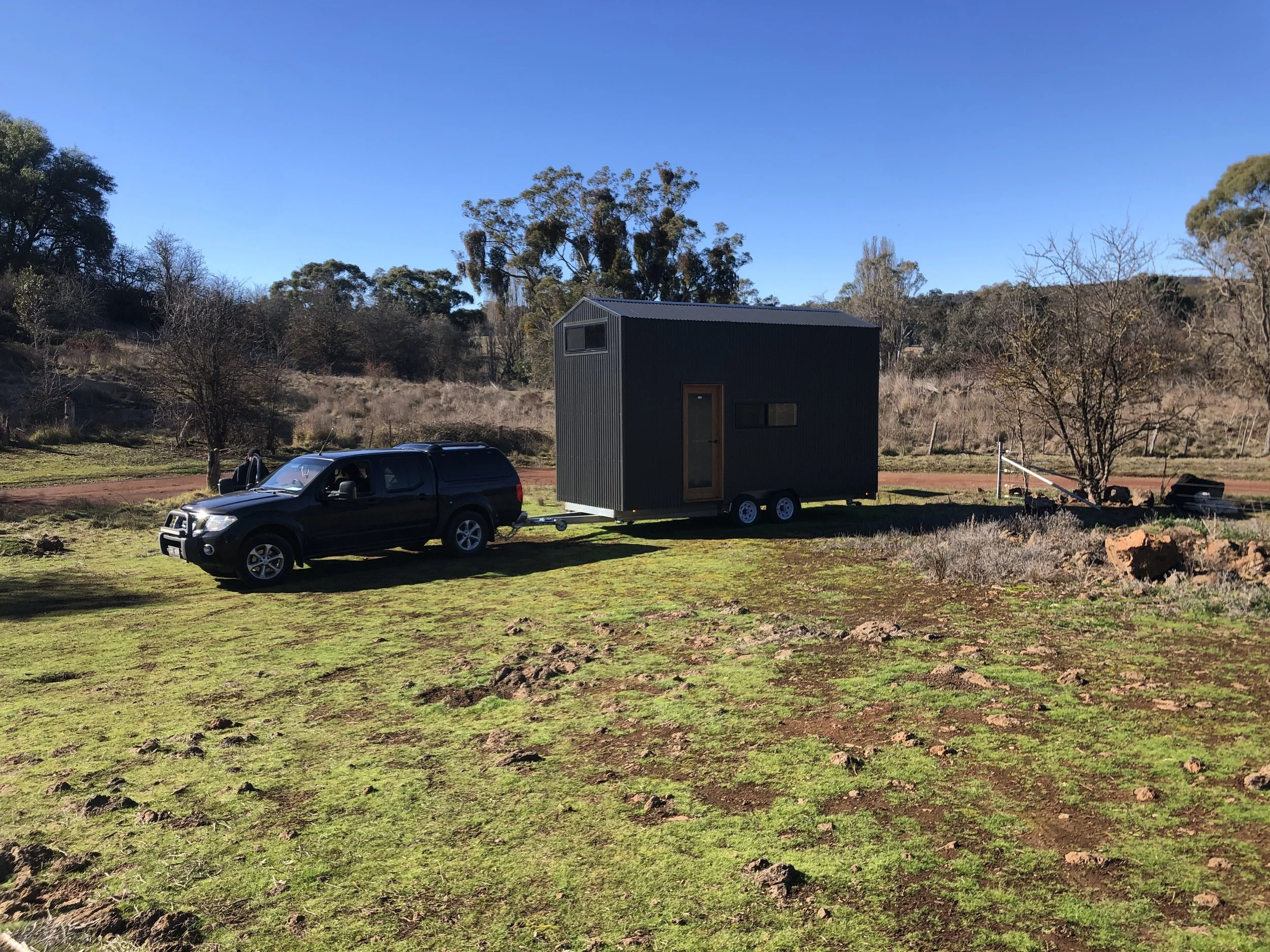 A black pickup truck pulling a tiny black modern house on wheels across a grassy area with a dirt road, surrounded by trees and hills under a clear blue sky.