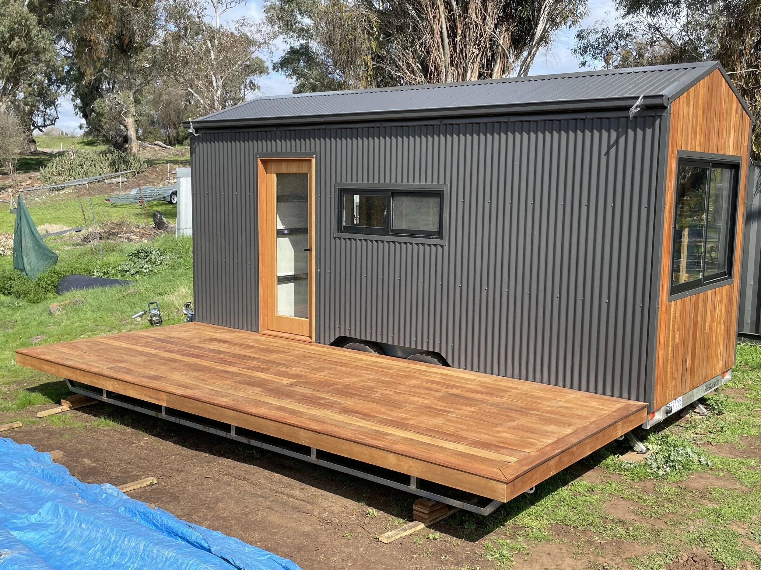 A small modern trailer with black corrugated metal walls and wooden accents, featuring a glass door and windows, sits on a raised wooden deck outside on grass and dirt.