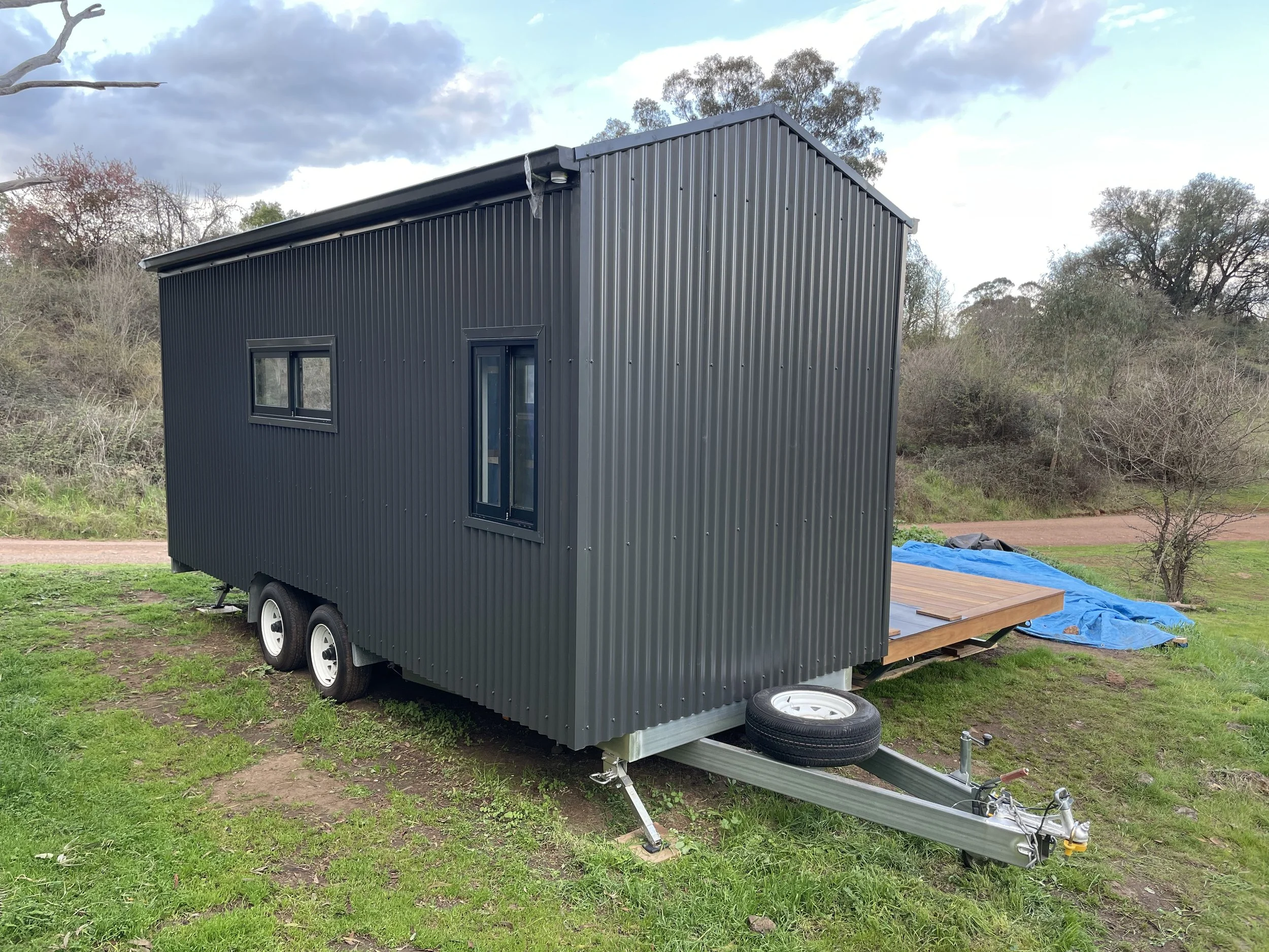 A small, black, metal tiny house on wheels with a wooden deck and two black framed windows, parked outdoors on grassy ground with trees and a dirt road in the background.