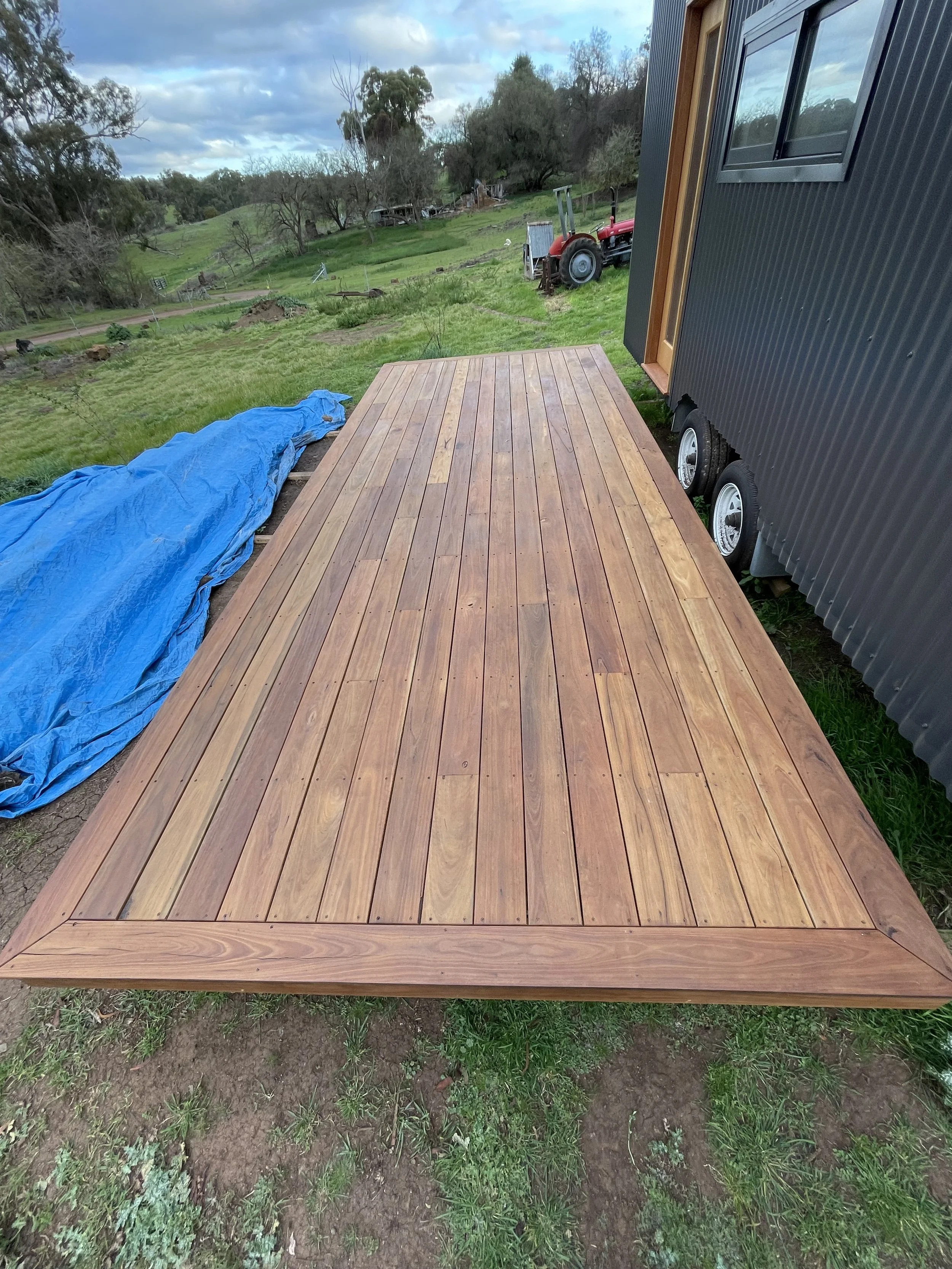 Newly installed wooden deck in a rural area with a blue tarp on the ground, a small red tractor, and a metal building with a window to the side.