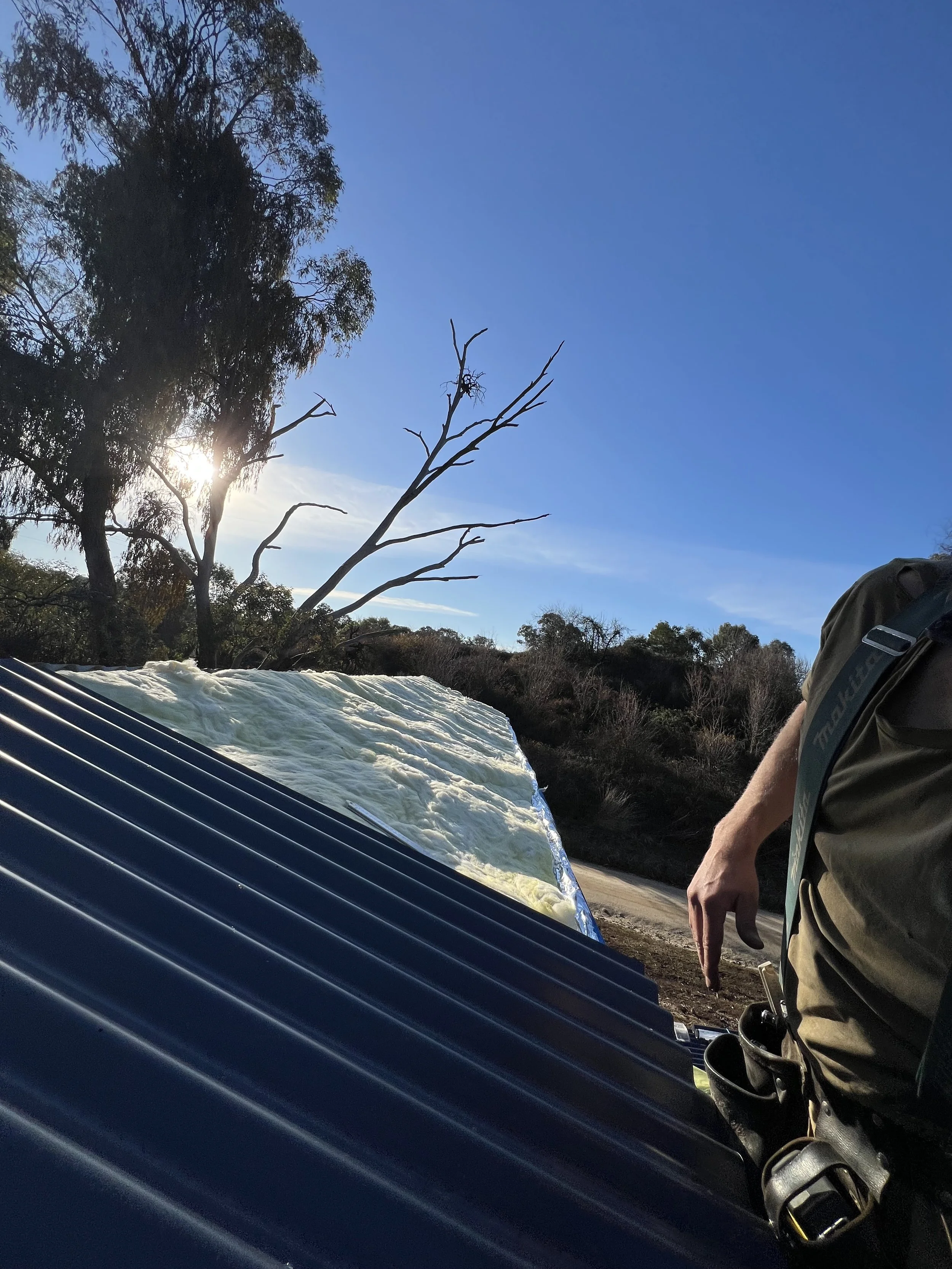 Person standing outdoors next to a solar water heater with a black corrugated surface and a reflective surface, under a clear blue sky with the sun partially obscured behind a tree, surrounded by trees and brush.