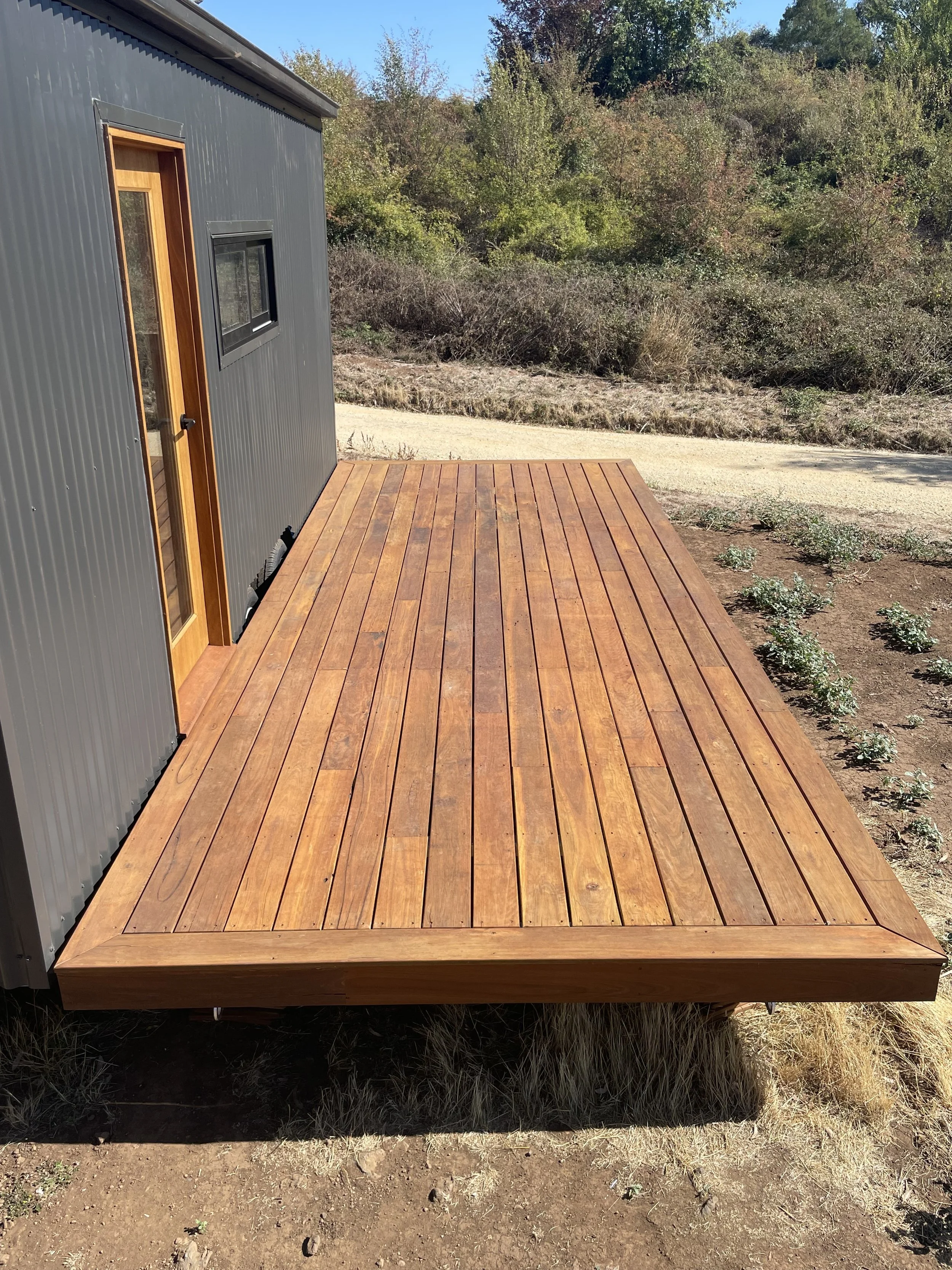 Wooden deck attached to a modern house with gray exterior siding and a wooden door, overlooking a landscape with dry grass, small plants, trees, and a dirt pathway.