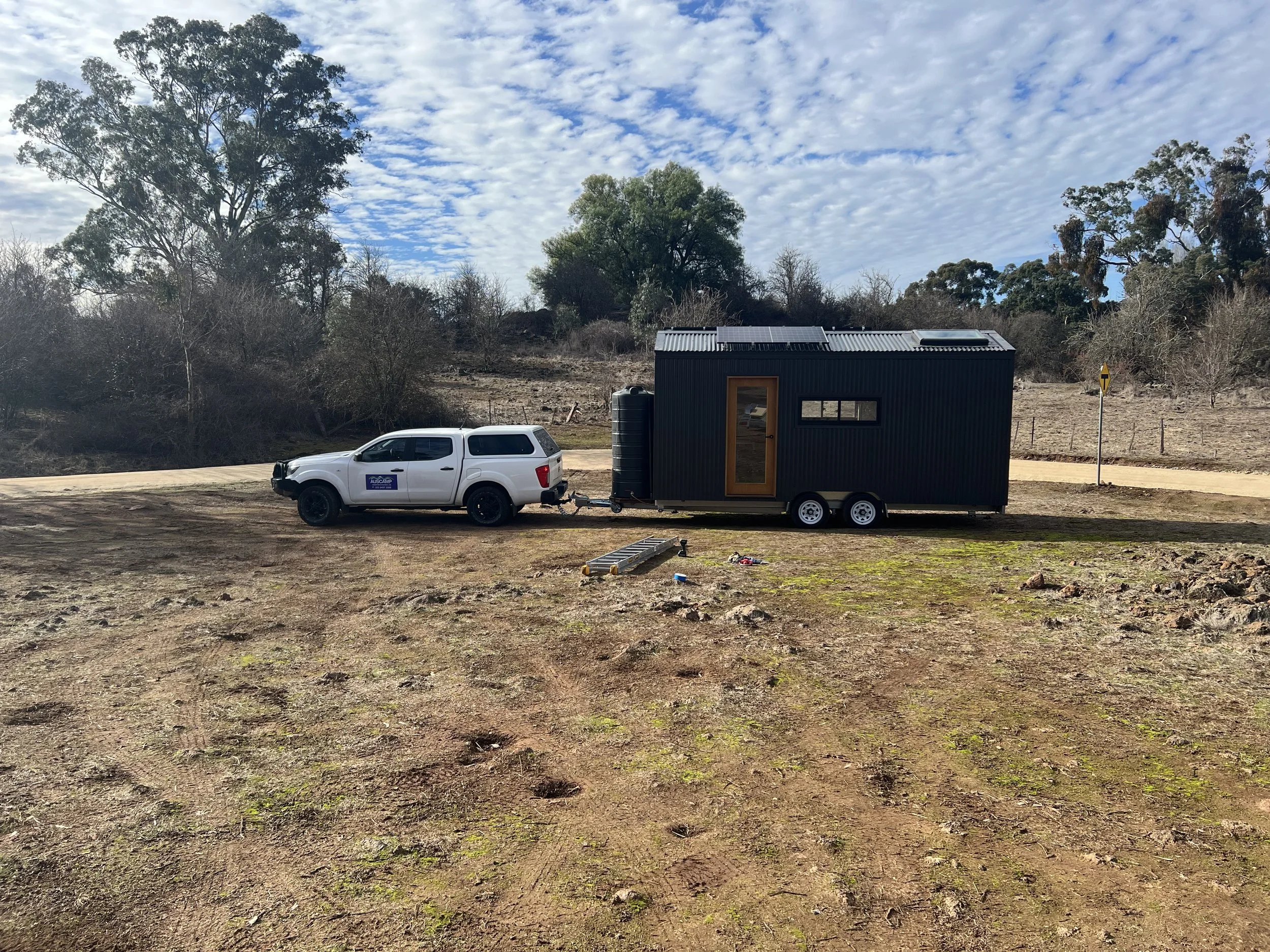 A white ute towing a black tiny house on wheels, parked on a dirt area with trees and a partly cloudy sky in the background.