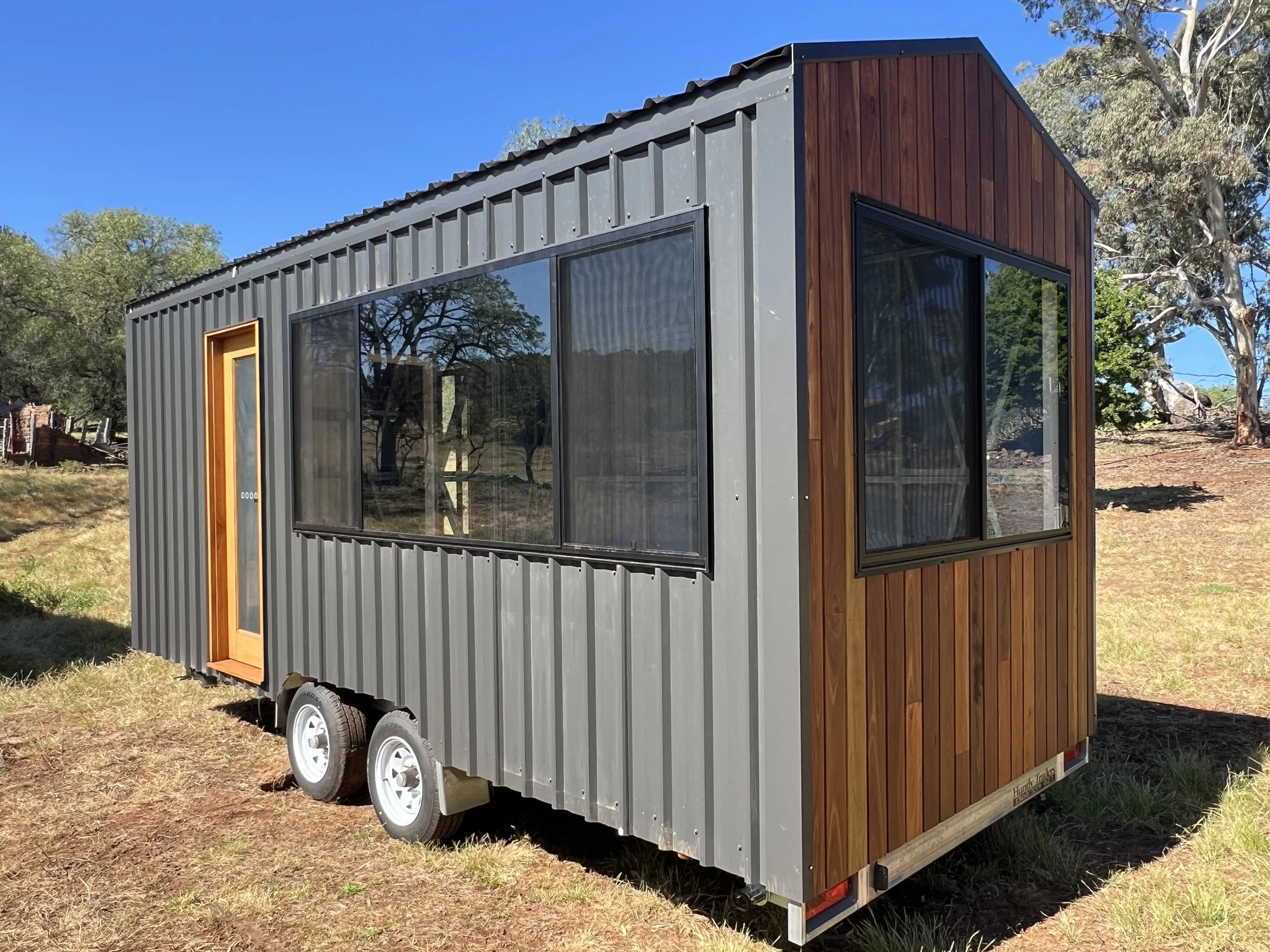 A small tiny house on wheels with gray metal siding and wood accents, situated on a grassy area with trees in the background.