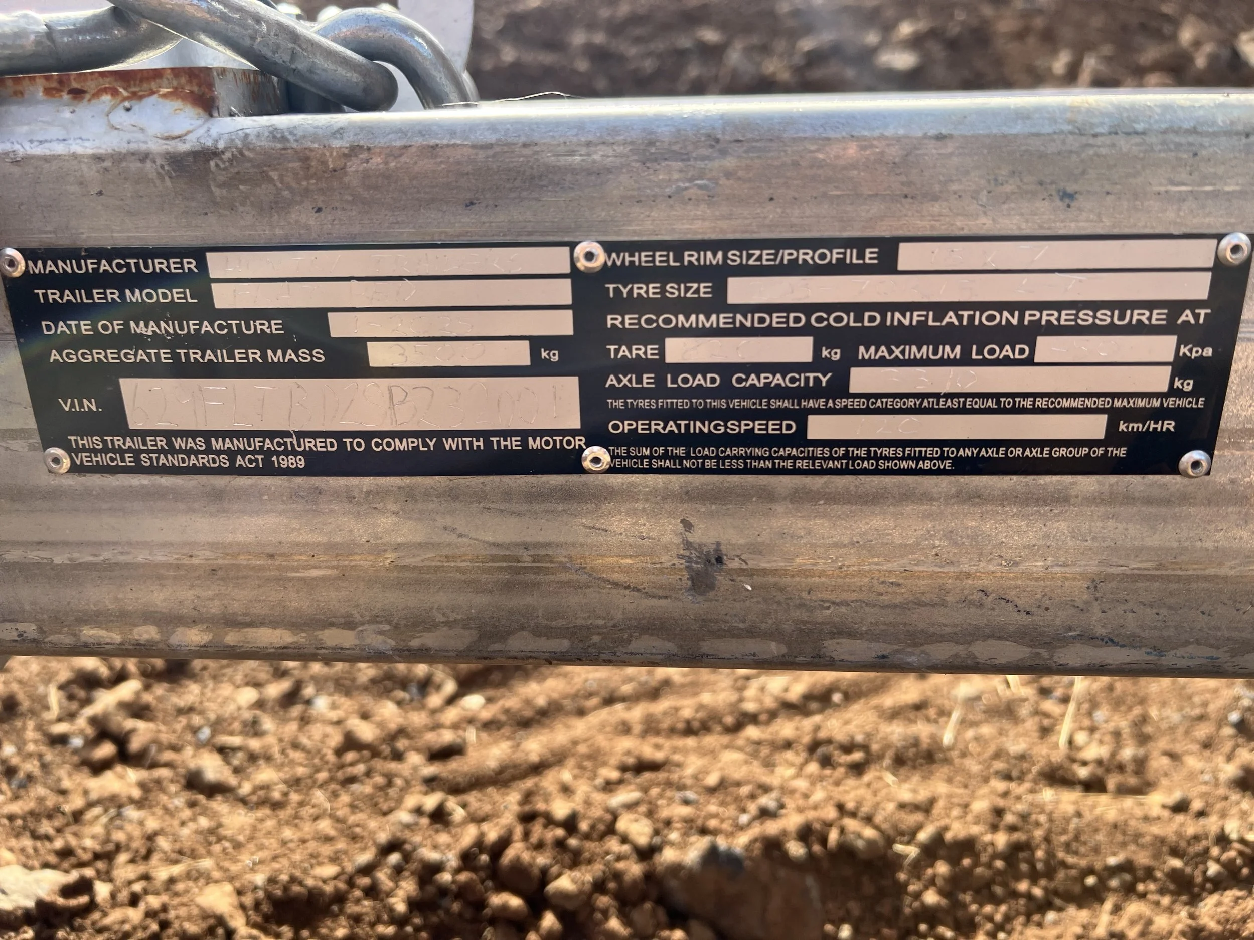 A metal trailer identification plate with fields for manufacturer, trailer model, date of manufacture, aggregate trailer mass, VIN, wheel rim size, tire size, cold inflation pressure, maximum load, axle load capacity, operating speed, and a statement