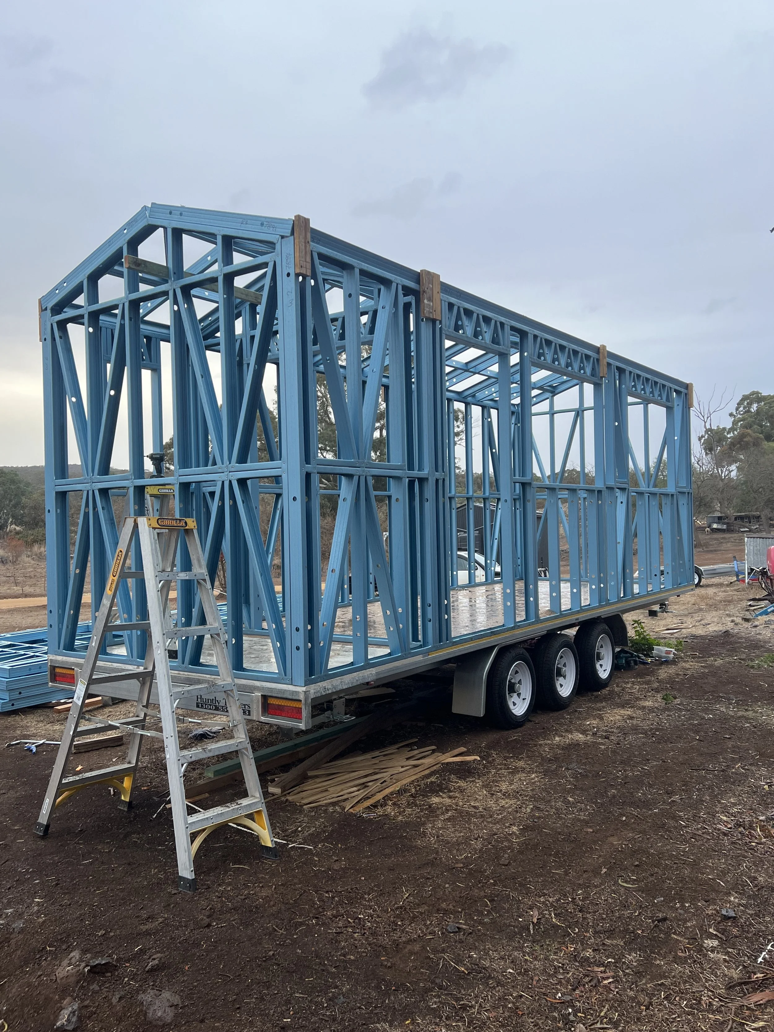 Blue steel framing on a trailer, with a ladder leaning against it, located outdoors on a dirt surface.