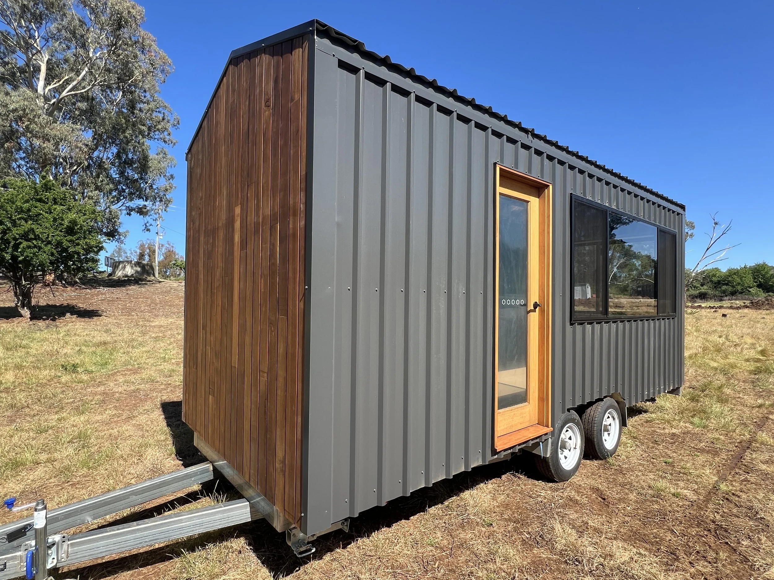 A tiny house on wheels with monument trimdek cladding, wooden door, and large windows, situated outdoors on a grassy area with trees in the background.