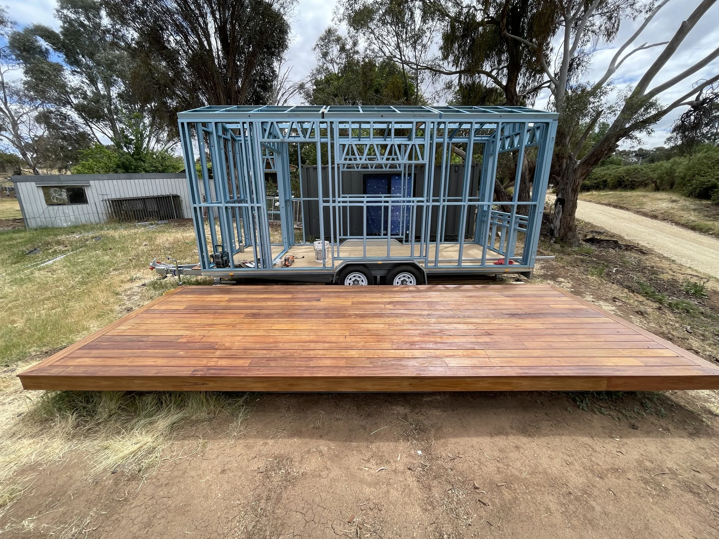 A construction site with a wooden deck in the foreground and a blue metal house frame on a trailer in the background, surrounded by trees and a dirt road.