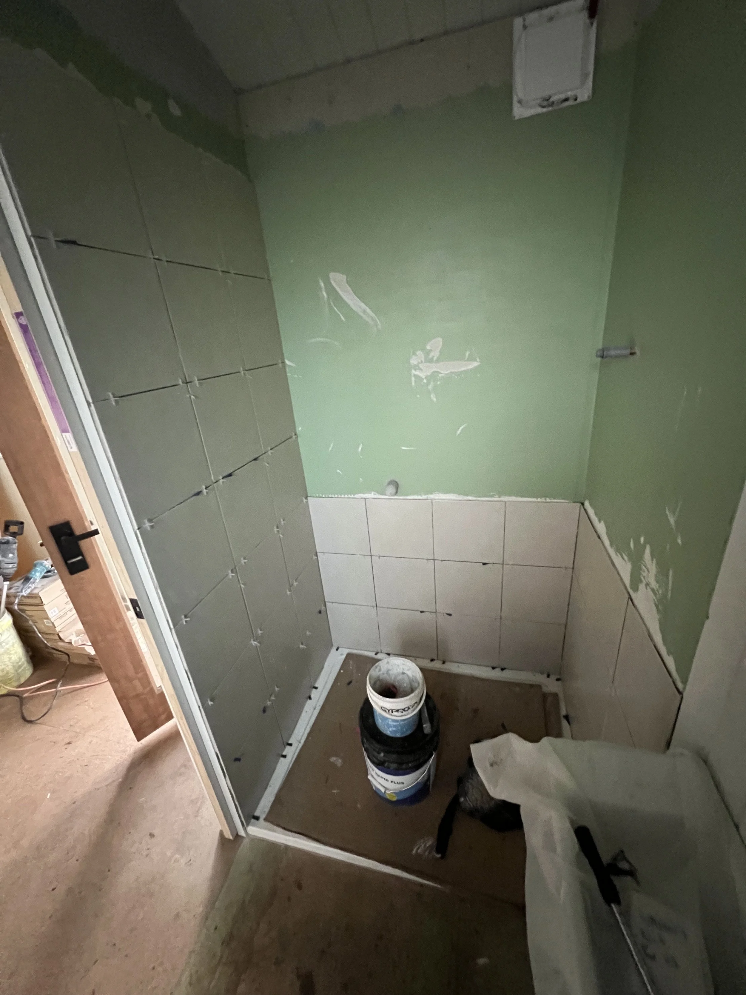 Small bathroom or shower room under construction with tiled walls, waterproofed walls, and construction materials including a bucket and a plastic bag on the floor.