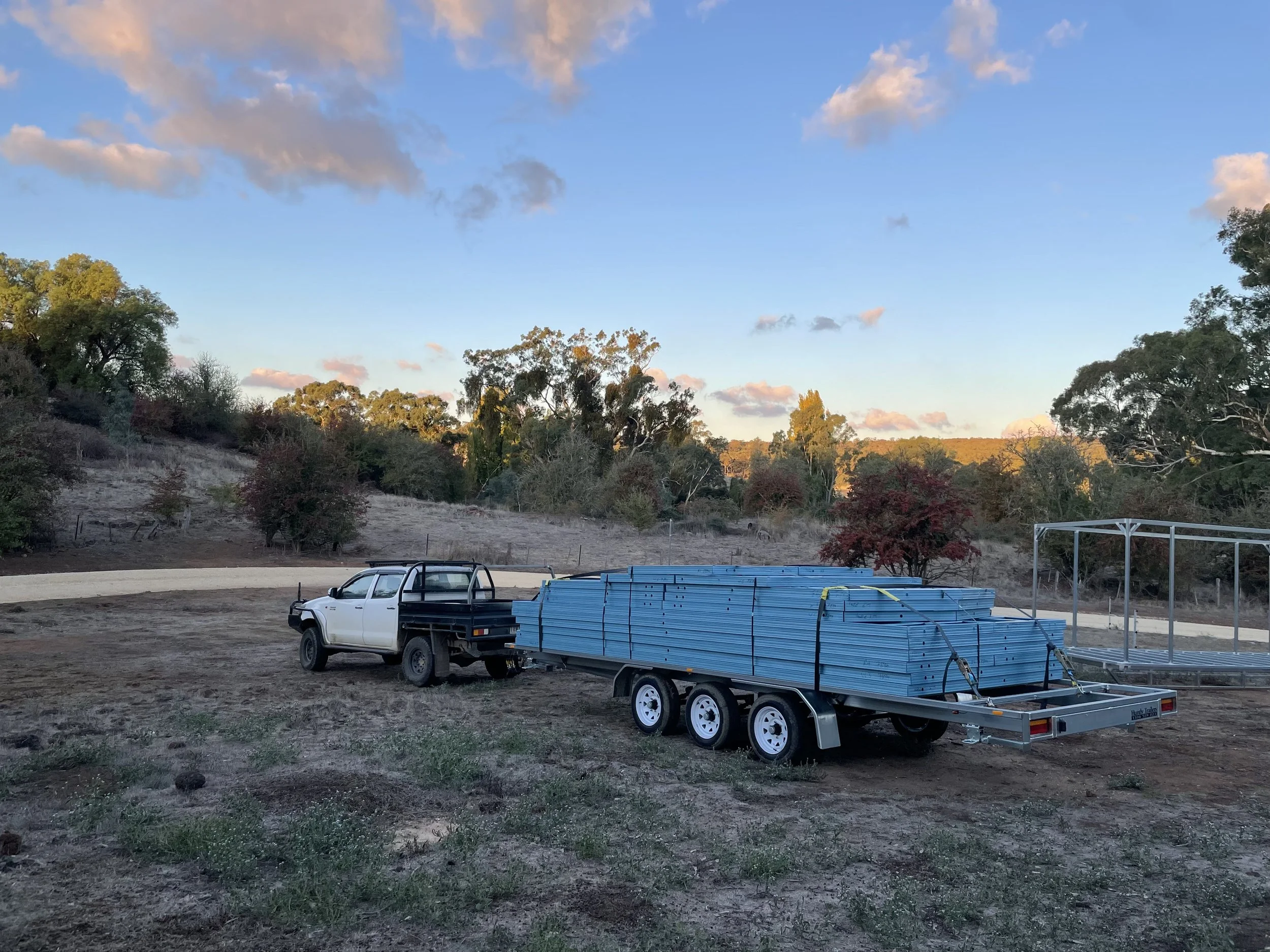 A white pickup truck with a black flatbed trailer attached, carrying blue metal poles, parked on a dirt area near a curved paved road, with trees and a partly cloudy sky in the background during sunset.