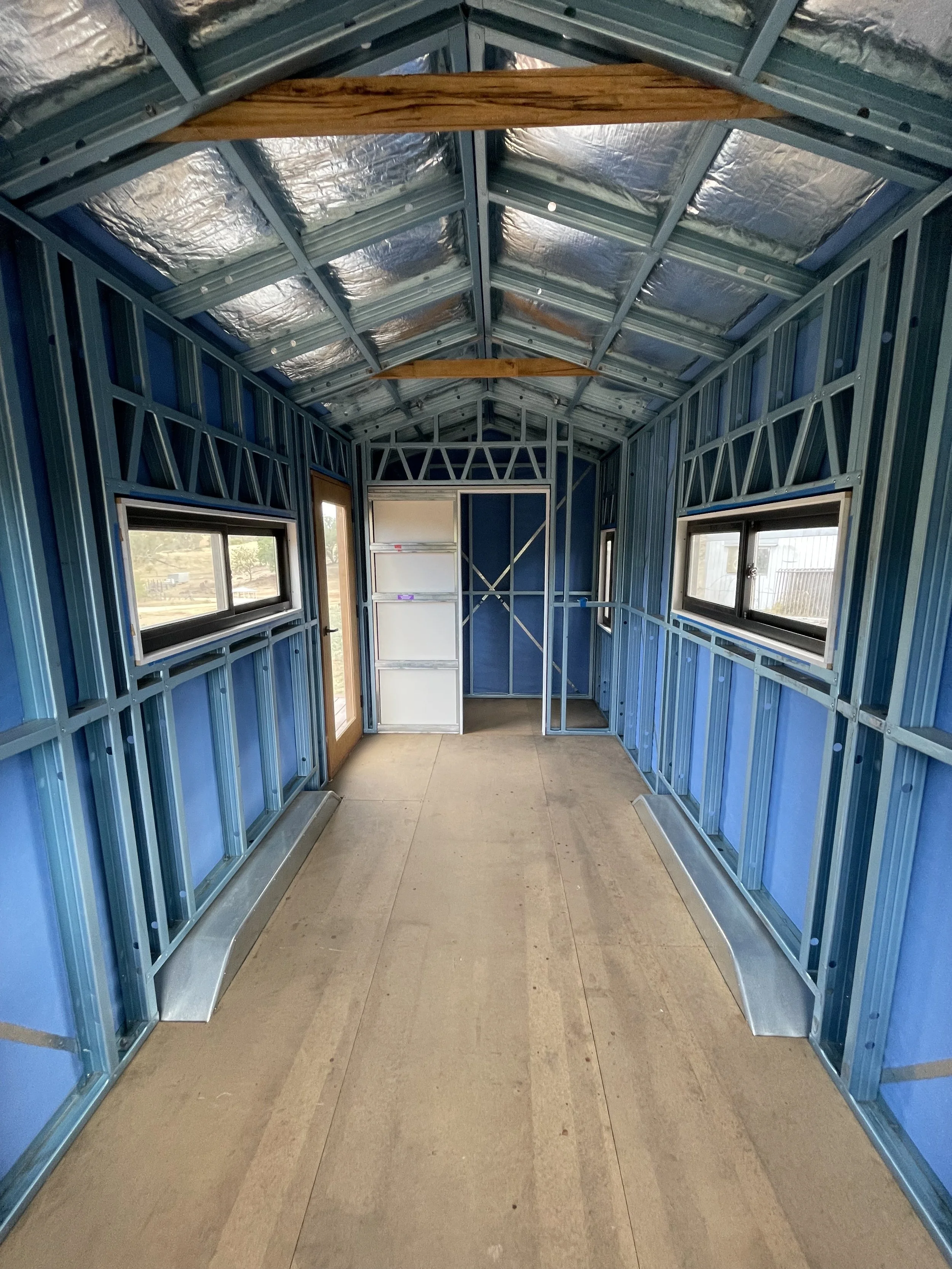 Interior of a small room under construction with blue metal framing, two rectangular windows, a door, and a sloped ceiling with insulation and wooden beams.