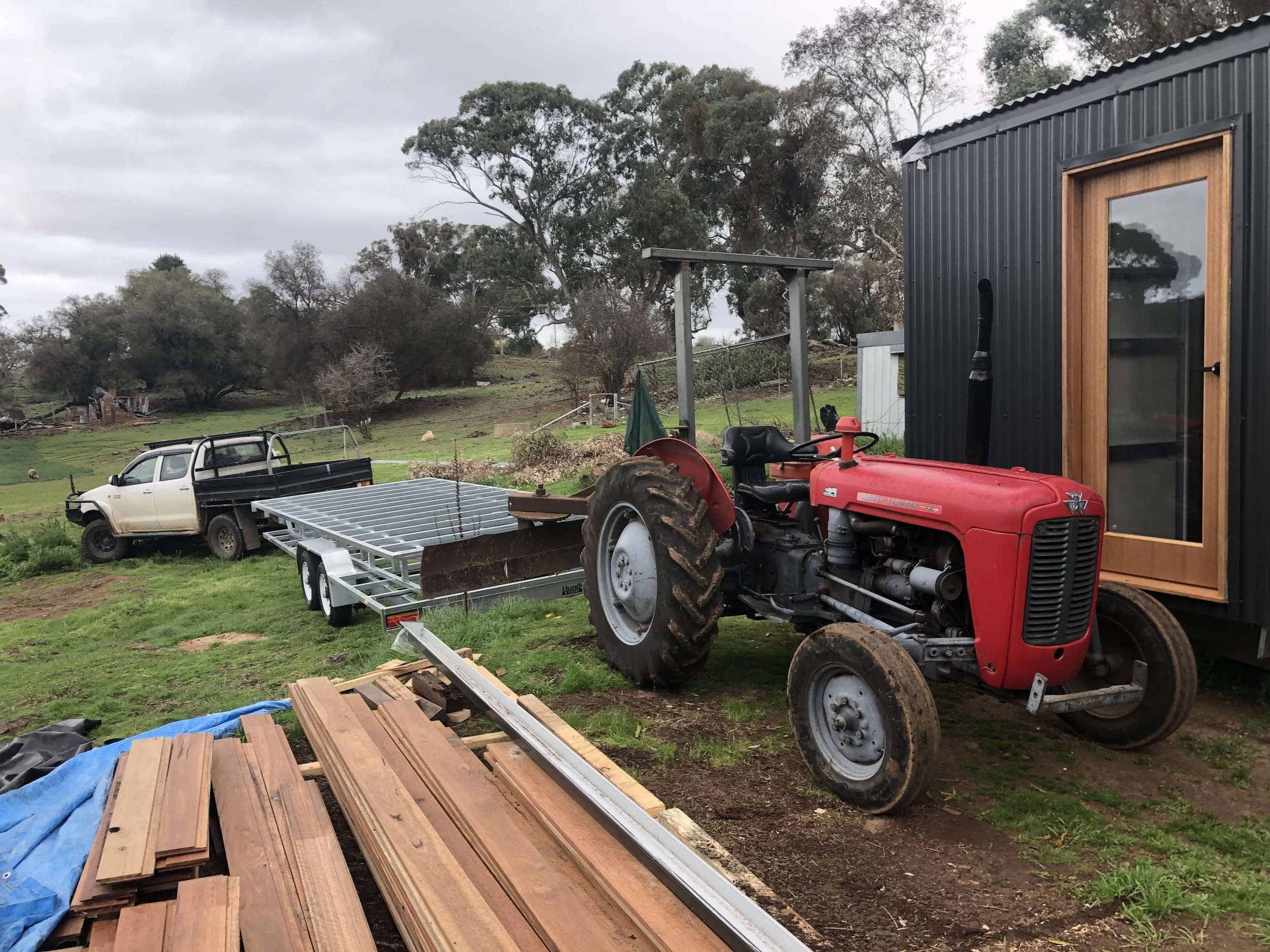 Farm scene with a red tractor, a black building with wooden window frame, a trailer, a white pickup truck, and surrounding rural landscape with trees and cloudy sky.