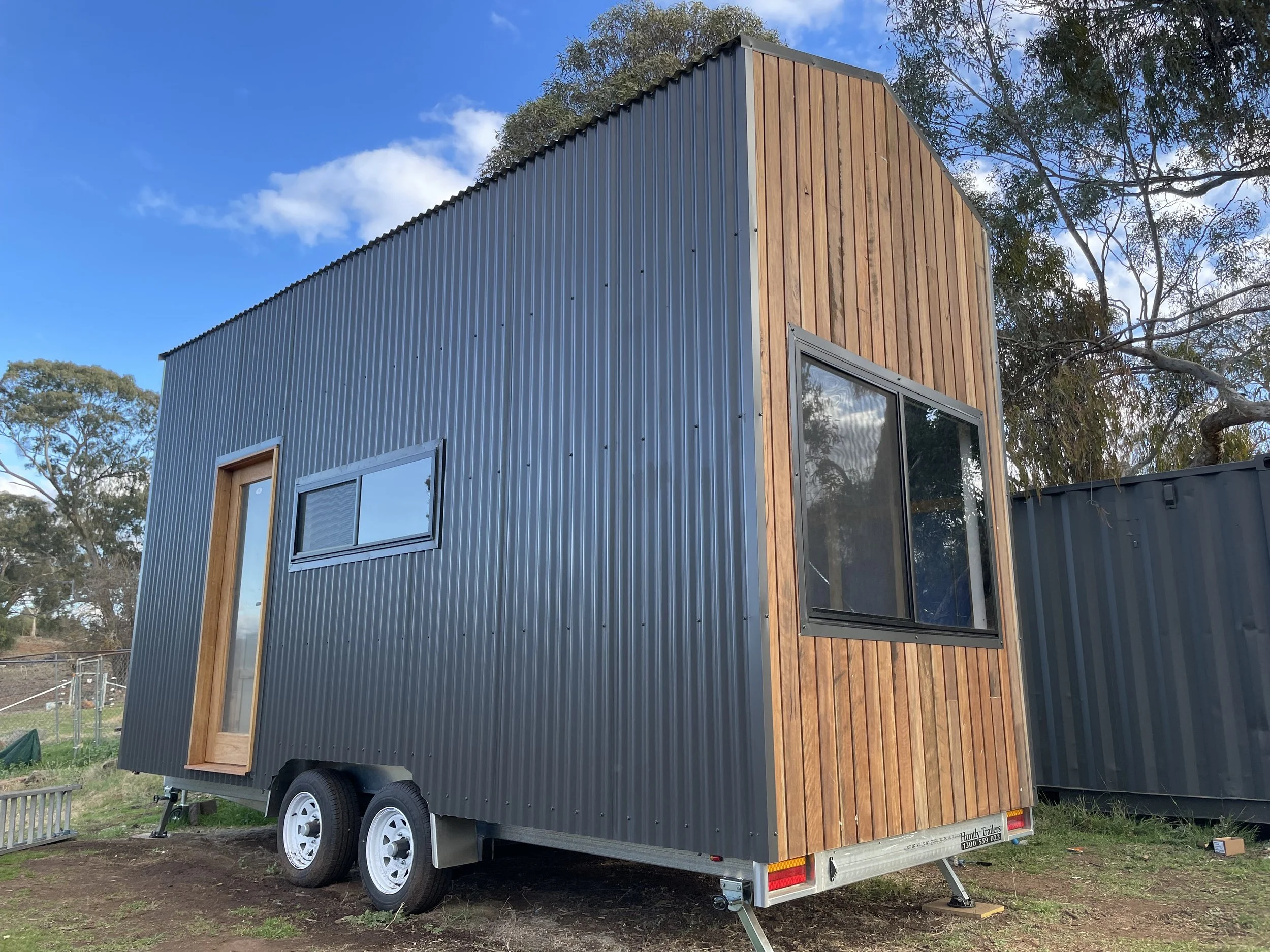A tiny house on wheels with metal siding and wooden accents, situated on a grassy area under a partly cloudy sky.