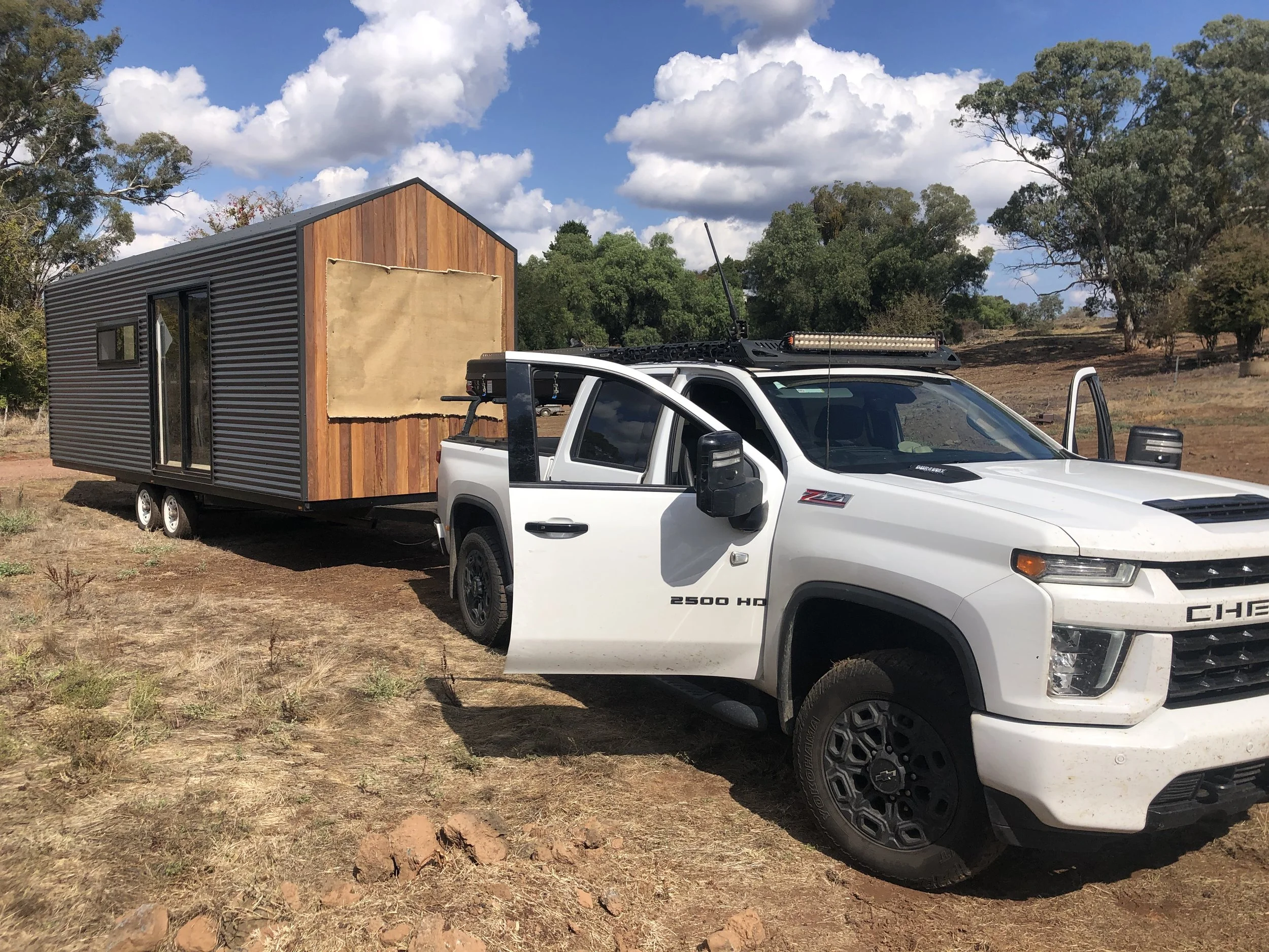 A white pickup truck pulling a tiny house trailer in a rural area with trees and clouds in the sky.