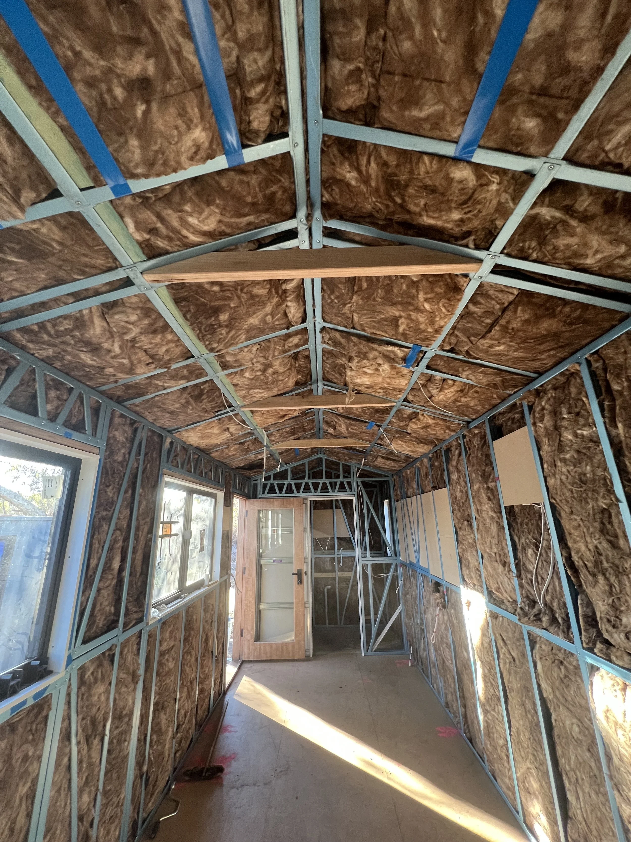 Interior of a room under construction with metal framing, insulation, windows, and a door.