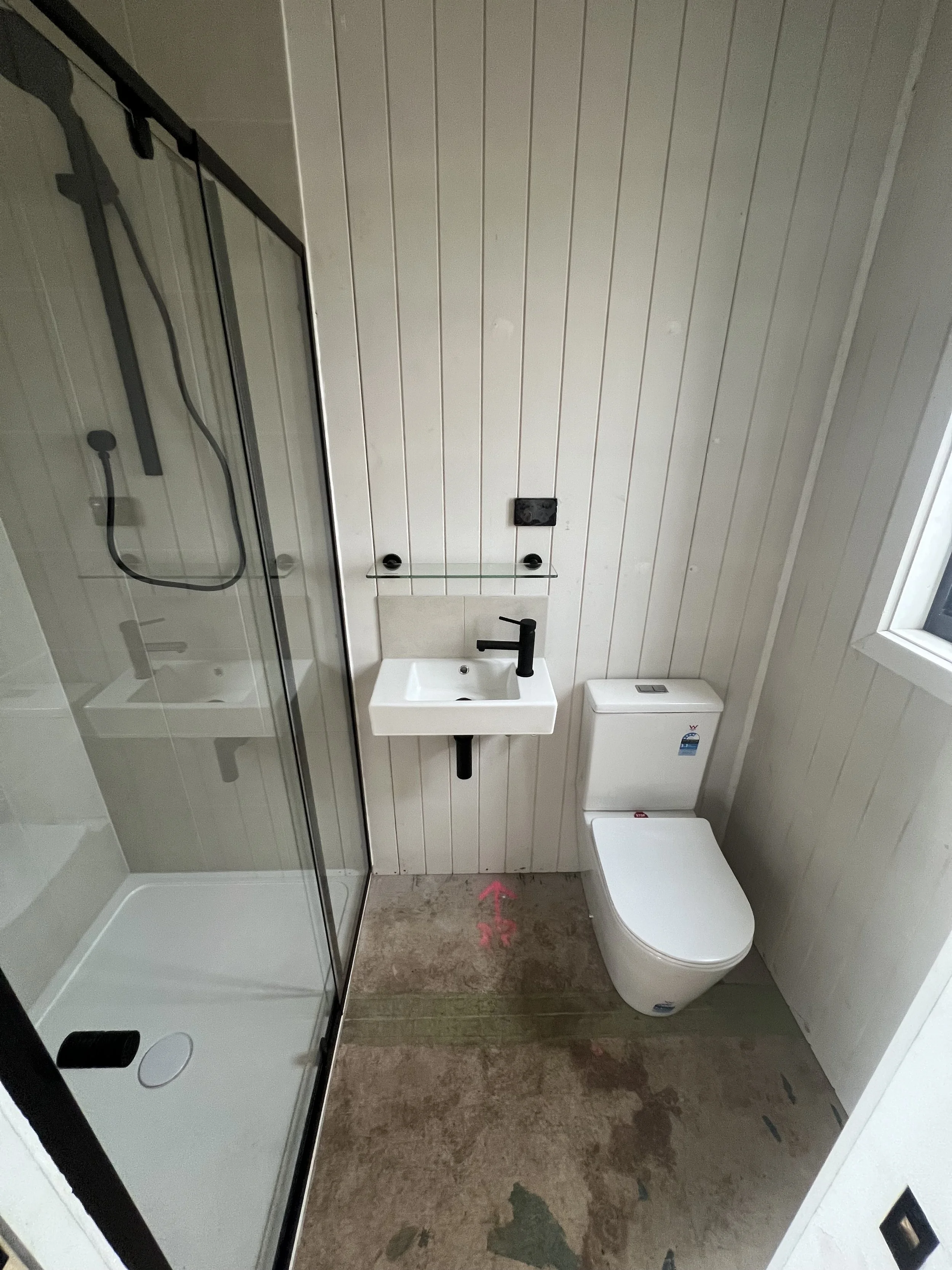 Small bathroom with a corner shower, small wall-mounted sink with black faucet, toilet, and a window, with unfinished flooring and white paneled walls.