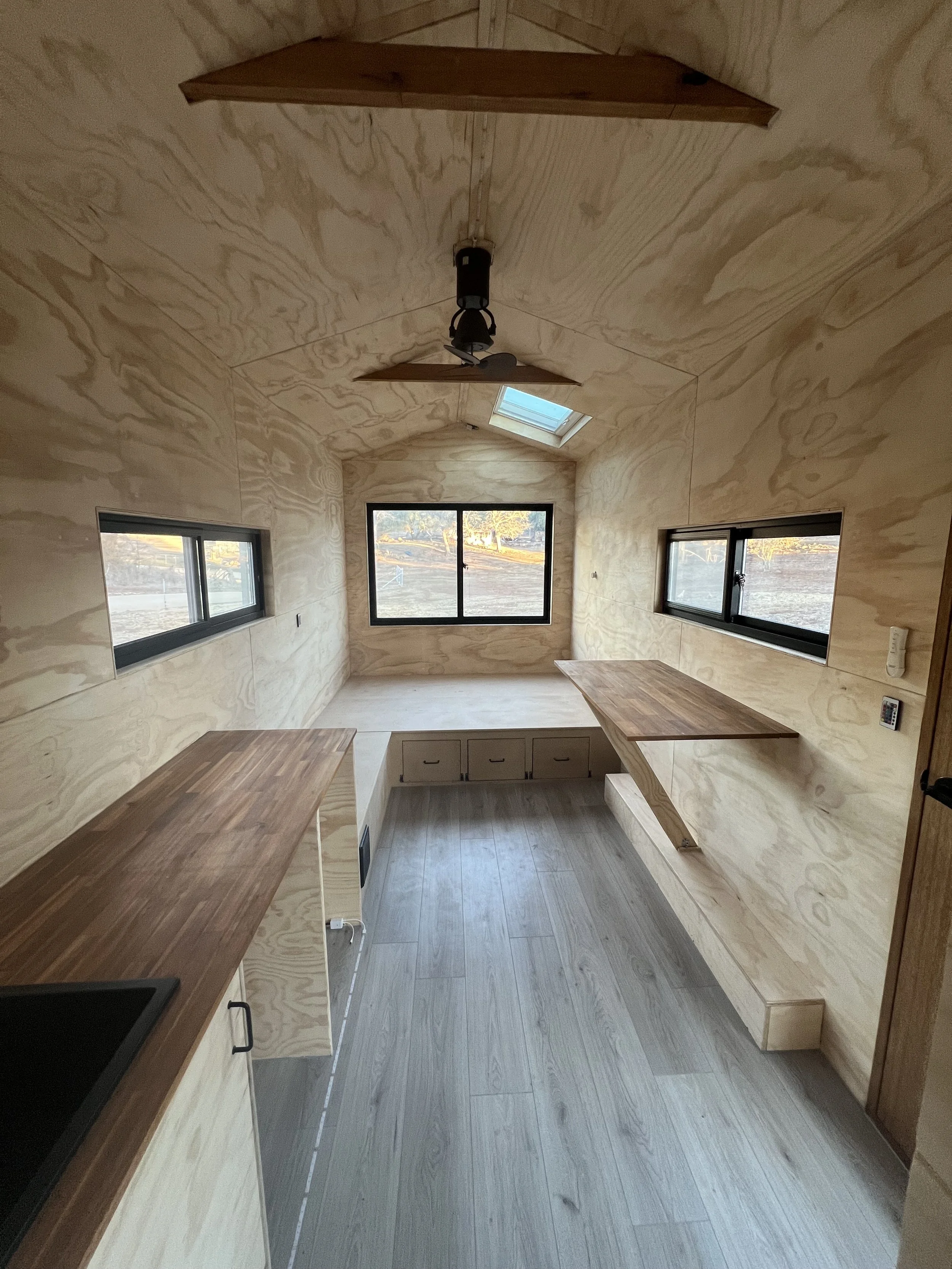 Interior of a small, unfinished room with plywood walls and ceiling, a skylight, three windows, two wooden shelves, and a wooden countertop with drawers underneath.