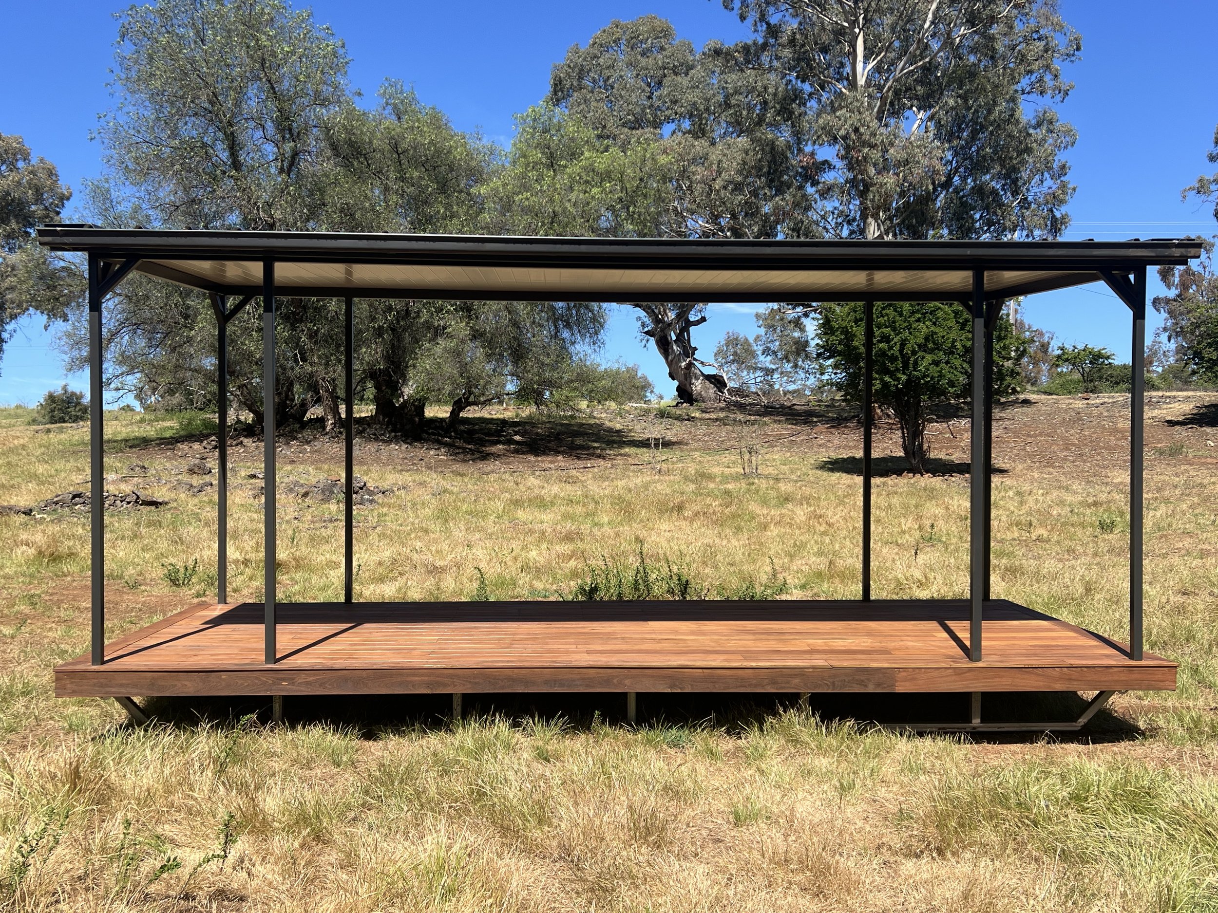 A metal and wood outdoor deck with a roof, situated in a grassy field with trees and clear blue sky.