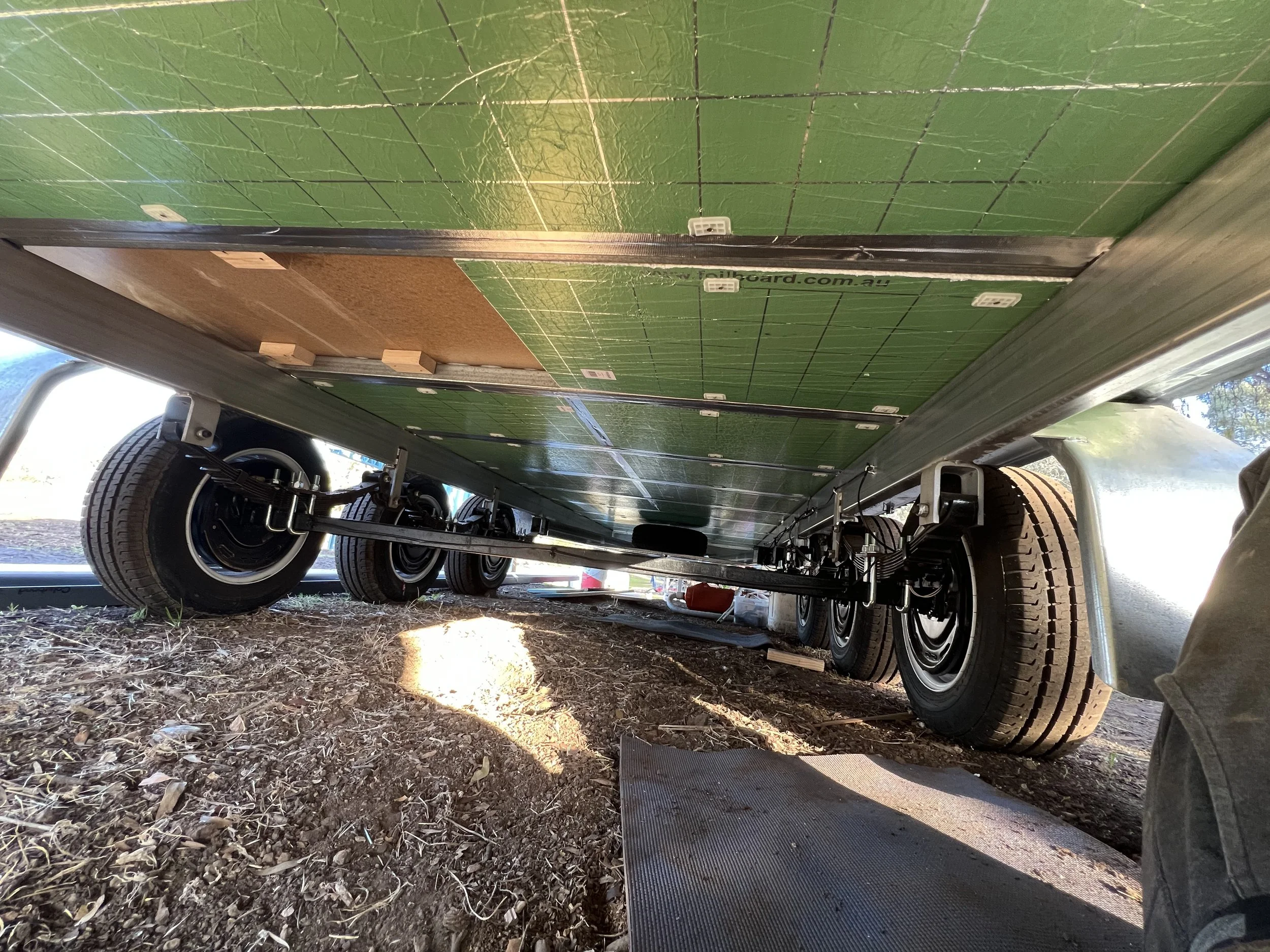 Underneath view of a mobile home or trailer with six wheels visible, situated on dirt ground with some leaves and a mat, with sunlight shining from the left side.