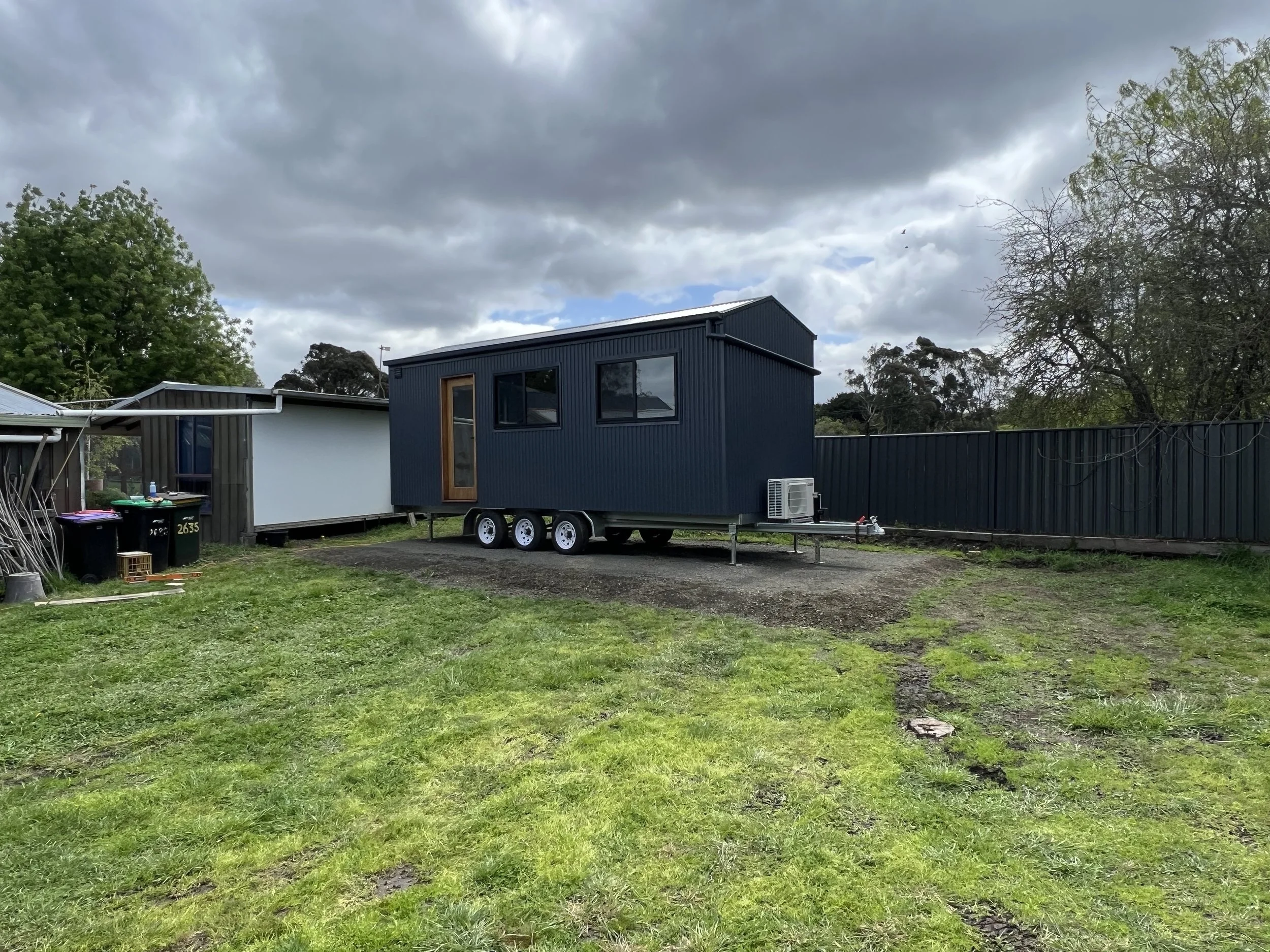 A tiny house on wheels painted dark blue with black windows and a brown door, situated in a backyard with green grass and a gravel area. There are some trees, a metal fence, and a cloudy sky in the background.